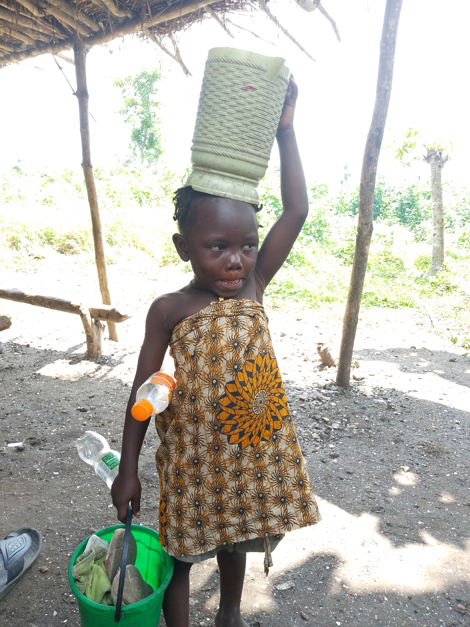 A young child, likely of African descent, stands outdoors beneath a thatched roof structure. The child wears a patterned cloth around their waist and holds a light green, woven container on their head. In their right hand, they grip a green bucket containing what appears to be cloth and other items. To their left, a plastic water bottle with an orange cap is attached to their arm. A second, empty plastic water bottle lies on the ground near the child's left side. The ground is covered in dirt, gravel, and scattered debris. In the background, lush green foliage and trees are visible, suggesting a rural or semi-rural environment. The lighting indicates it is daytime with strong sunlight. The location is identified as Otulu, Nigeria.