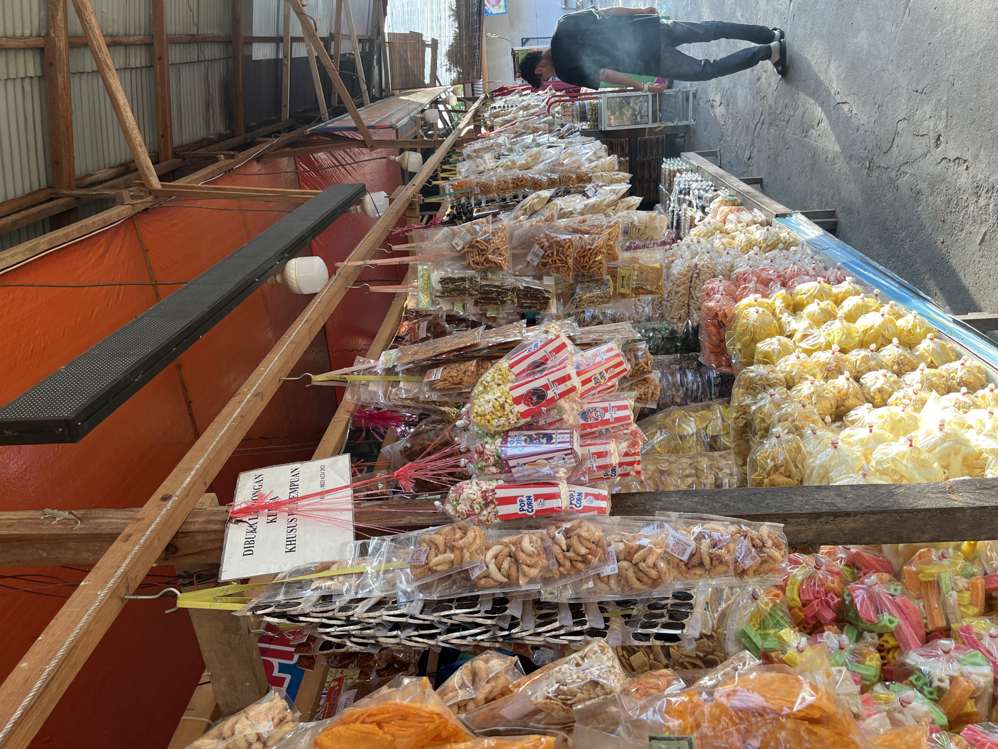 The image depicts an outdoor market stall in Sawang, Indonesia, where a variety of snacks are displayed for sale. The stall is covered by a corrugated metal roof and an orange tarp. Wooden beams and posts support the structure, with various items hanging from hooks along a wooden beam. These hanging items include numerous individually packaged snacks such as popcorn in red and white striped bags, various fried snacks, and what appear to be dried or processed food items.
In the foreground, a long wooden table is laden with more packaged snacks. These include colorful, star-shaped crackers in the front left, followed by rows of yellow and white puffed snacks, and then various bags of fried snacks and other packaged goods. A person, appearing to be a man wearing a black t-shirt and blue jeans, is walking away from the stall towards the right side of the image, casting a long shadow on the concrete ground. The ground appears to be a paved surface, possibly a sidewalk or market floor.
A sign with text in Indonesian is visible hanging from a hook. The text on the sign reads: "DIBUKAKAN KITA KHUSUS PEREMPUAN" which translates to "We Open For Women Only". Below this, a phone number is partially visible. The overall scene suggests a busy, local market environment.