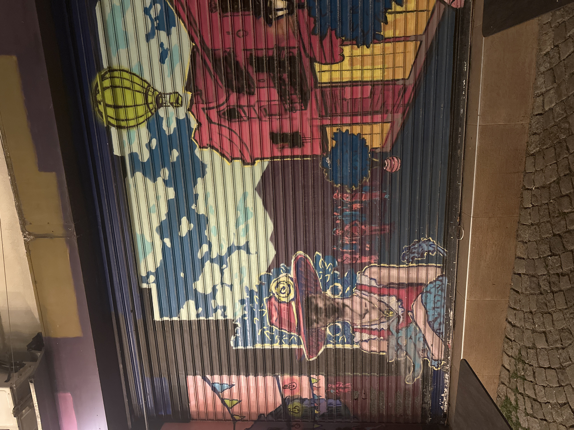 This image depicts a vibrant graffiti mural covering a closed metal roller shutter in Plovdiv, Bulgaria. The artwork features a surreal street scene with a central figure wearing a wide-brimmed red hat adorned with a yellow rose. The figure is dressed in a red top with suspenders and appears to be holding something in their lap.
Behind the figure, the mural depicts stylized pink buildings with yellow accents and a dark mountain range under a blue sky dotted with clouds and a yellow hot air balloon. The roller shutter itself is made of horizontal metal slats, which are partially visible through the paint, giving the image a textured appearance.
The mural is located at street level, with a cobblestone pavement visible in the foreground. The overall style of the graffiti is bold and colorful, with a distinct cartoonish aesthetic. The lighting suggests it is evening or night, as artificial light casts shadows on the pavement. The roller shutter appears to be on the storefront of a commercial establishment.