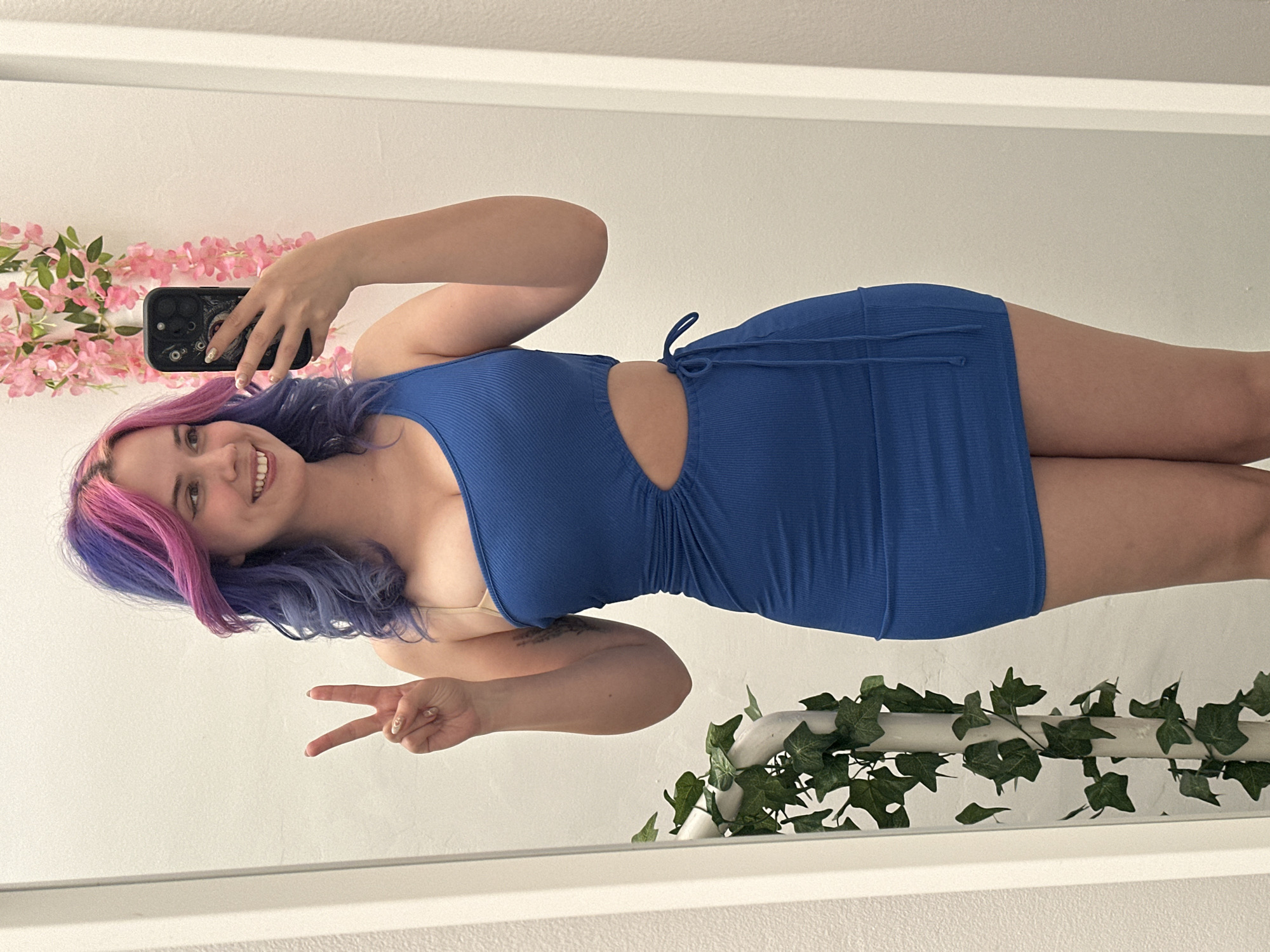A young woman is captured in a mirror selfie, posing with a wide smile and a peace sign made with her right hand. The image is taken indoors, likely in a well-lit room with white walls, and is reflected in the mirror.
The woman has long, wavy hair, dyed a vibrant blue/purple with notable pink streaks at the front and top. She is wearing a form-fitting, ribbed, royal blue mini-dress that features a cutout on the left side of her waist, revealing skin, and a drawstring detail on the right side of her hip. A light-colored bra strap is visible on her left shoulder. Her nails appear to be painted with a light color and possibly a design. In her left hand, she holds a black smartphone with a detailed case, suggesting she is actively taking the photo. A faint tattoo is partially visible on her right forearm.
The background, reflected in the mirror, shows a white wall adorned with decorative elements. On the left side, a pink floral garland with green leaves is draped horizontally. On the right side, a green ivy garland is similarly arranged, appearing to rest on a ledge or frame. The overall atmosphere conveyed by her expression and pose is happy and playful. While not visually confirmed in the image, the provided context indicates the location is the City of Zagreb, Croatia. No discernible text is visible on any objects in the image.