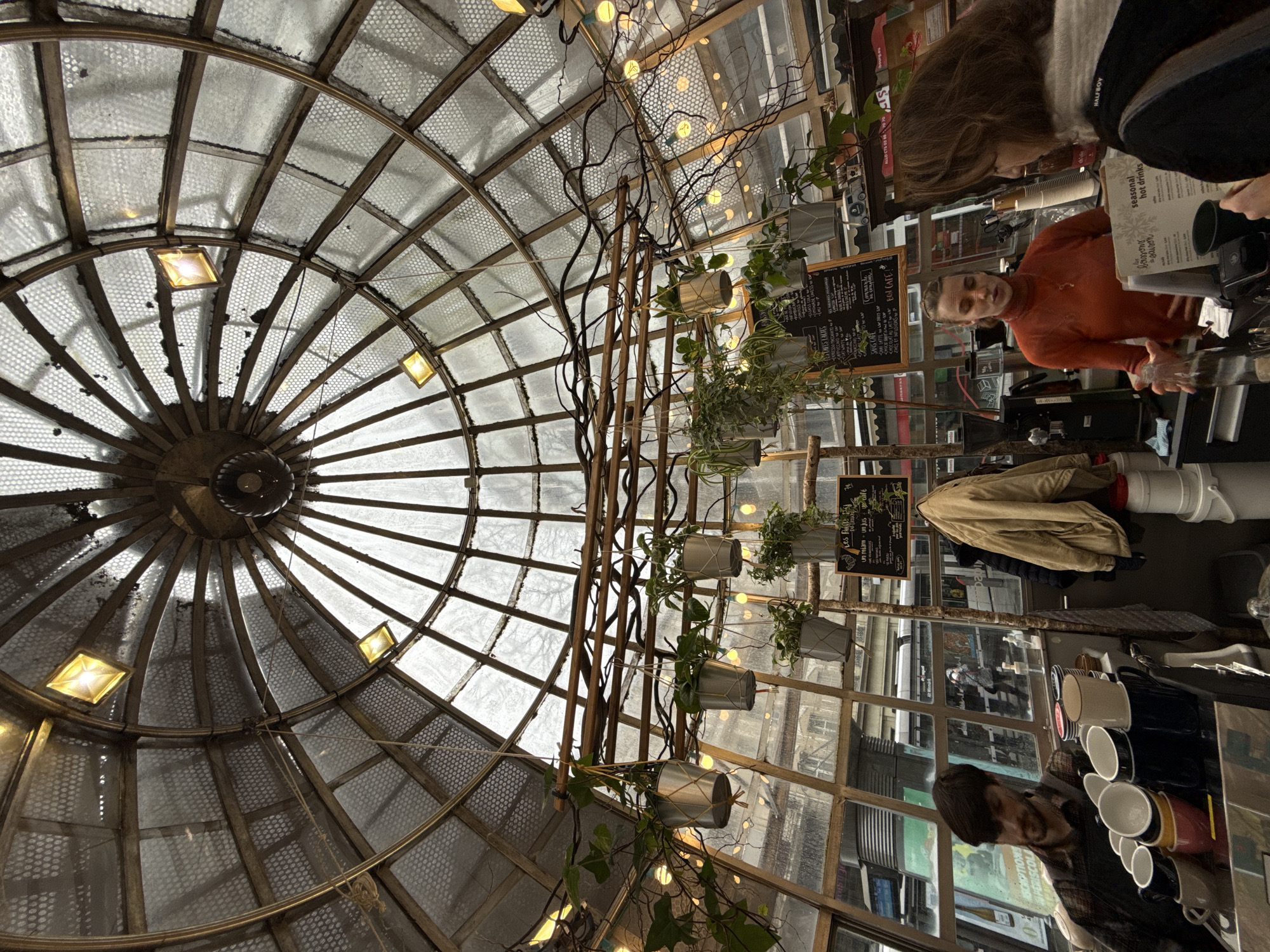 The image captures the interior of a glass-domed cafe or kiosk, identified as being located in Strasbourg, France. The perspective is looking upwards and forwards, showcasing the domed ceiling structure and the service area below.
The ceiling consists of a radial metal framework supporting multiple panels; some are clear glass, allowing diffused natural light, while others appear to be perforated metal or frosted glass, creating a textured pattern. Several square, warm-toned light fixtures are integrated into the dome's framework. Beneath the dome, an arrangement of bare branches and numerous hanging plants in small silver pots, suspended by ropes, forms a decorative canopy. String lights with circular, warm-glowing bulbs are interwoven within this greenery.
At counter level, three individuals are present. On the far left, a male with dark hair and a beard, wearing a plaid shirt, is partially visible behind a counter that holds a stack of multi-colored ceramic cups and other beverage preparation equipment. In the center, a female employee with dark hair, wearing an orange long-sleeved top, stands behind the counter, gesturing with her right hand while holding a clear glass bottle in her left. She is engaged in an interaction with a female customer on the right, whose back and right profile are visible. The customer has brown hair, wears a dark jacket with a grey hooded element, and holds a white menu card titled "seasonal hot drinks."
Behind the employees, two chalkboard menus with handwritten French text are mounted. A beige jacket hangs on a hook to the right of the male employee's station, near a stack of white buckets. Through the clear glass panels of the lower dome structure, a blurred urban background is visible, featuring a modern tram (likely a city tram) and indistinct reflections of buildings and street activity. Sanitizer bottles are visible on the counter surfaces.