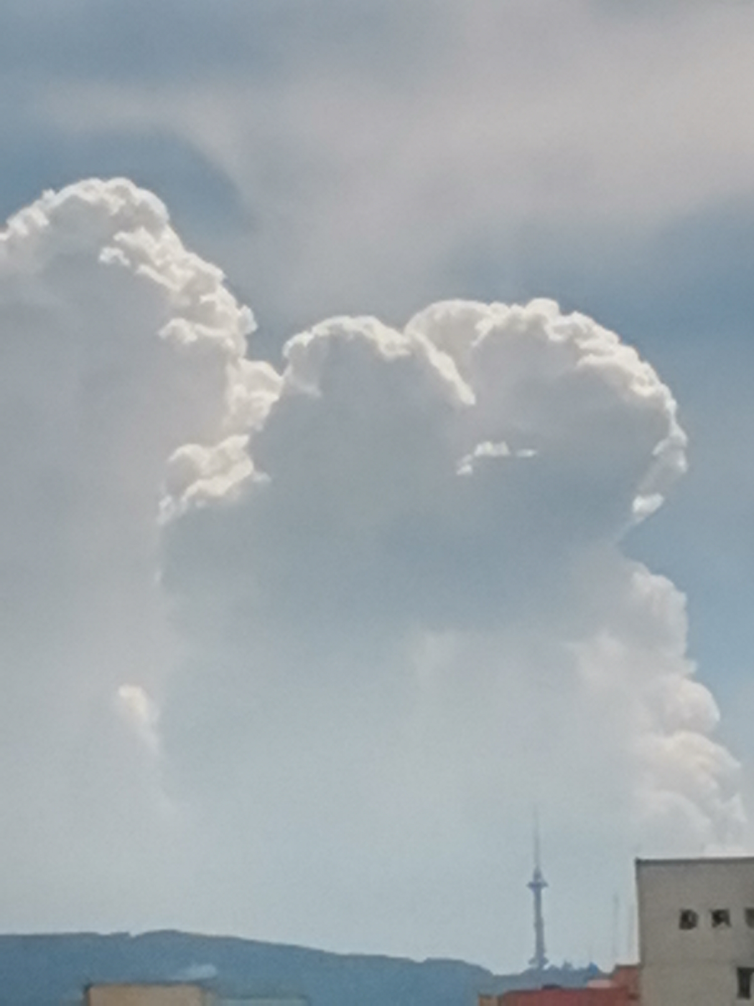 A wide shot captures a fluffy, white cloud formation in a bright sky, with hints of blue and gray. The cloud, which dominates the upper portion of the frame, appears to be shaped like a face, with discernible features. In the lower section of the image, a cityscape is visible, featuring a tall, slender tower and a building with windows. The tower is likely the Bulgarian National Television Tower, a prominent landmark in Sofia, Bulgaria. The overall scene is typical of a partly cloudy day in an urban environment.