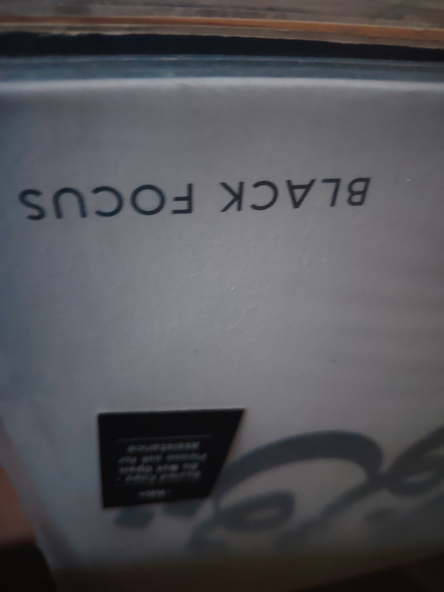 The image is a close-up, angled shot of what appears to be a light gray album or record cover. The item is positioned upside down relative to the viewer.
The most prominent feature is the bold, dark gray text "BLACK FOCUS" printed horizontally across the upper half of the cover. Since the item is inverted, "BLACK" appears on the right side and "FOCUS" on the left.
Below this text, on the lower left side of the cover, there is a small, dark rectangular label. The text on this label is also inverted and extremely blurry, making it largely illegible. However, some fragmented words like "assistance" and "Do Not Open" (or similar instructional phrases) might be partially discernible due to the structure of the letters.
To the right and below the small label, a faint, dark gray, abstract design or part of a graphic (possibly a stylized face or logo) is visible, partially obscured by shadows and blur.
The background at the very top of the image shows indistinct brown, orange, and dark lines, suggesting the presence of other items, possibly records or a shelf, above the primary subject. No people or specific activities are depicted. The overall setting appears to be indoors, but no further details about the location or time of day can be determined.