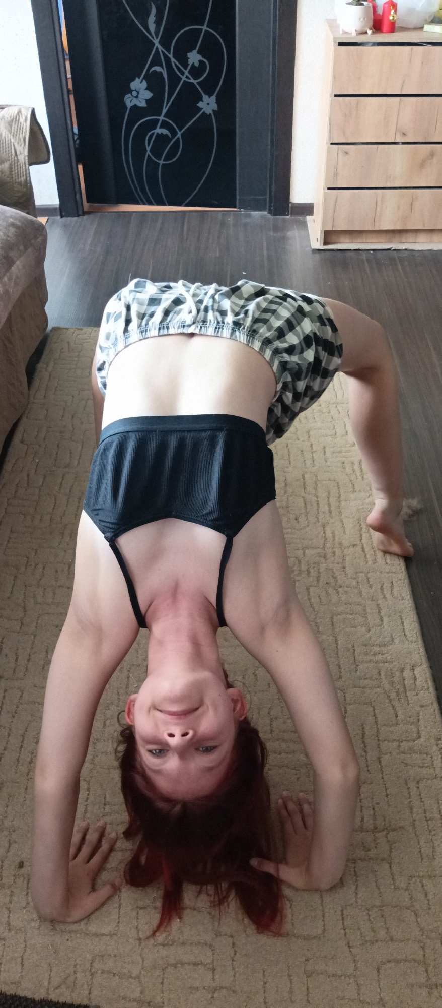 A person with auburn hair is in a backbend pose on a patterned tan rug, facing the camera. They are wearing a black top with thin straps and checkered shorts with a white drawstring. The background includes a wooden floor, a dark-colored door with a floral design, and a light-colored dresser with drawers. Part of a beige sofa is also visible on the left. The location appears to be indoors, possibly a residential space.
