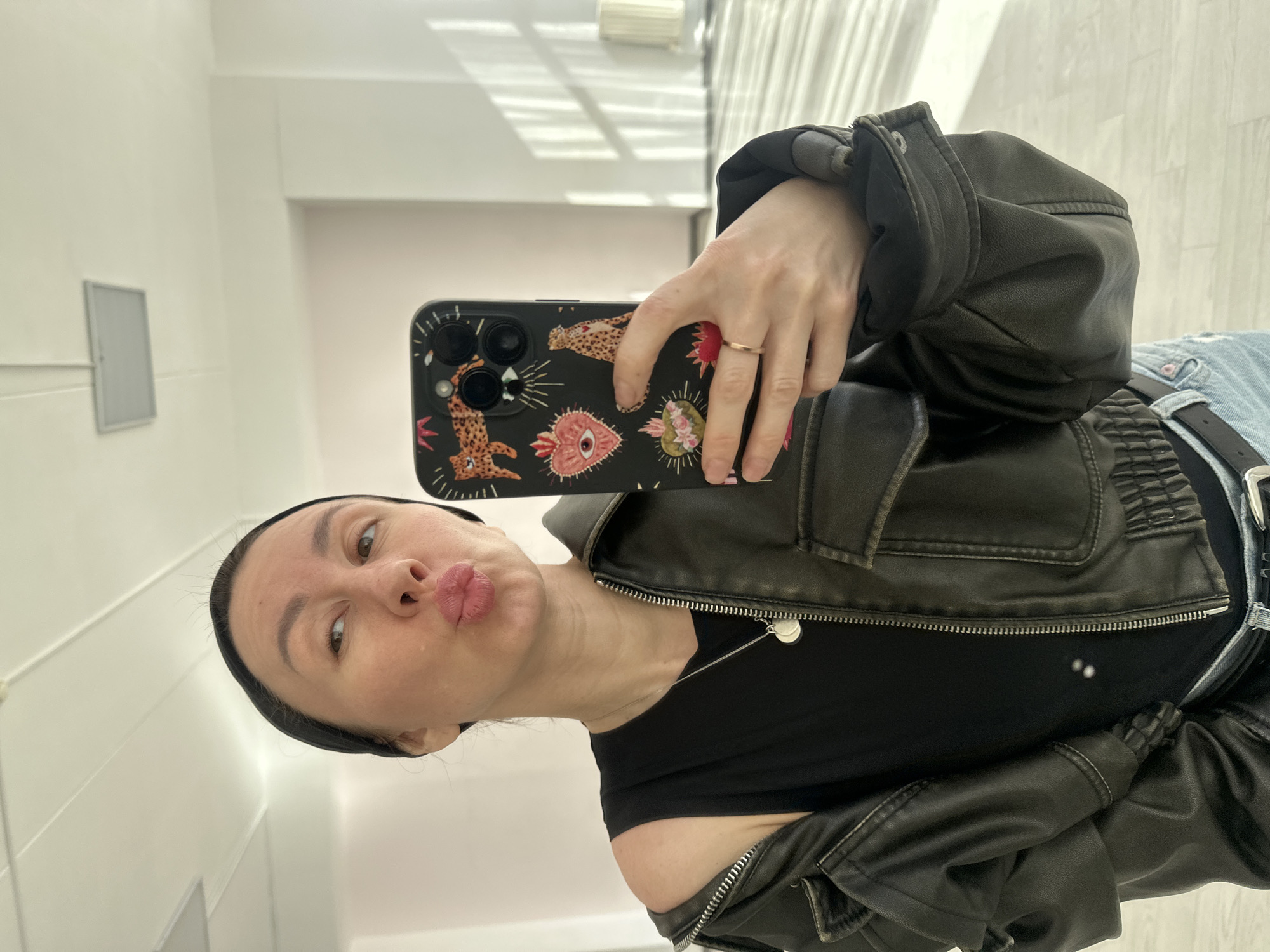 A woman is taking a selfie in what appears to be a well-lit interior space with white walls. She is wearing a black head band, a black t-shirt, and a dark leather jacket. She is pouting her lips for the photo. She is holding a black smartphone with a decorative patterned case. The background consists of white walls and a vertical window-like structure with light streaming through. The location does not appear to be in Votkinsk, Russia.