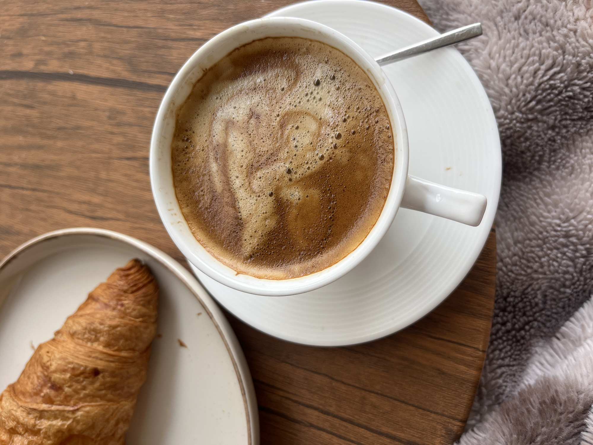 A close-up, high-angle shot shows a cup of coffee and a croissant on a wooden surface. The coffee cup, white and filled with a brown beverage, sits on a matching white saucer. A small silver spoon rests on the saucer near the cup. The coffee has a foamy surface. Beside the coffee cup is a croissant on a white plate with a slightly raised edge. Part of a soft, gray blanket or fabric is visible on the right side of the image, near the saucer. The wooden surface has a slightly glossy finish and appears to be a tabletop. The scene has a warm, inviting feel. The image does not provide any information about the location in the city of Budva, Montenegro.