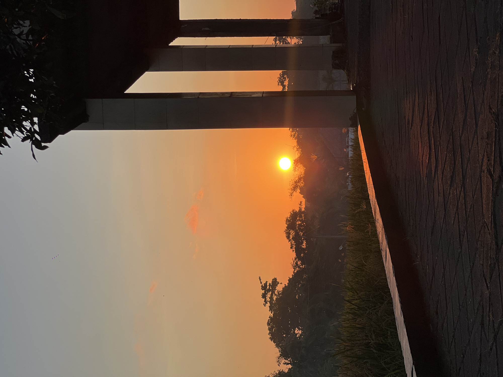 The image captures a vertical view of a sunrise in North Kuta, Indonesia. The sun is positioned amidst trees, casting an orange hue across the sky. The scene is partially framed by architectural elements, including pillars and a dark overhanging structure on the left and the corner of a walkway on the right. The walkway is paved with textured stones. Vegetation, including grass and trees, is visible along the ground. In the sky, a few dark, clustered shapes, potentially birds or kites, are seen on the upper left side.