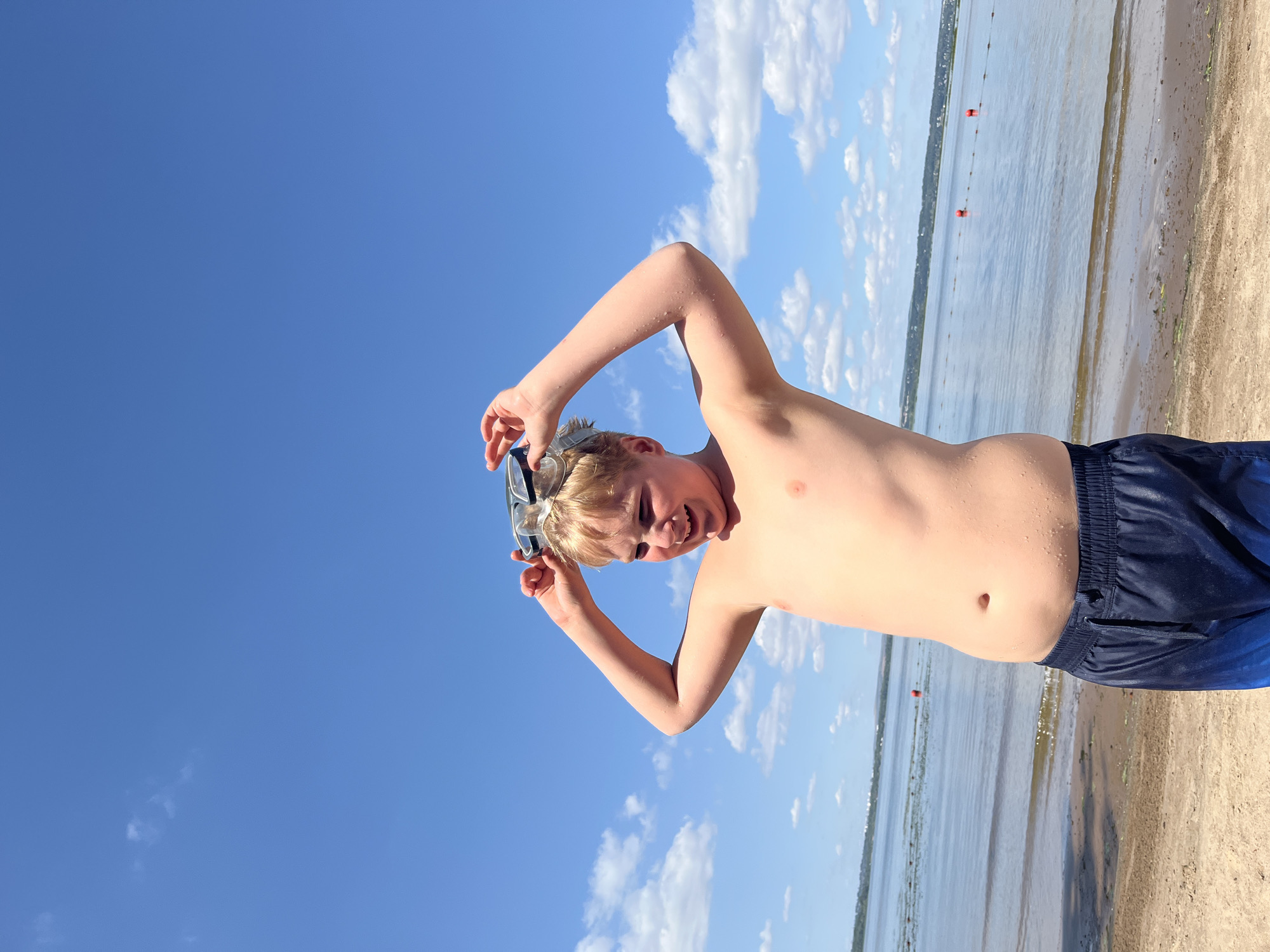 A young boy stands on a sandy beach, looking at the camera. He is shirtless and wearing dark blue swim trunks. The boy has wet hair and is holding a pair of goggles above his head. He has a wide smile on his face, suggesting he is happy and enjoying himself.
The background of the image shows a calm body of water, likely a lake or the sea, with a distant shoreline. There are a few buoys marking a swimming area in the water. The sky is a clear, bright blue with a few scattered white clouds, indicating fair weather and daytime. The sand on the beach appears light brown and is wet in some areas near the water's edge. The overall impression is of a sunny day at the beach, likely during summer.