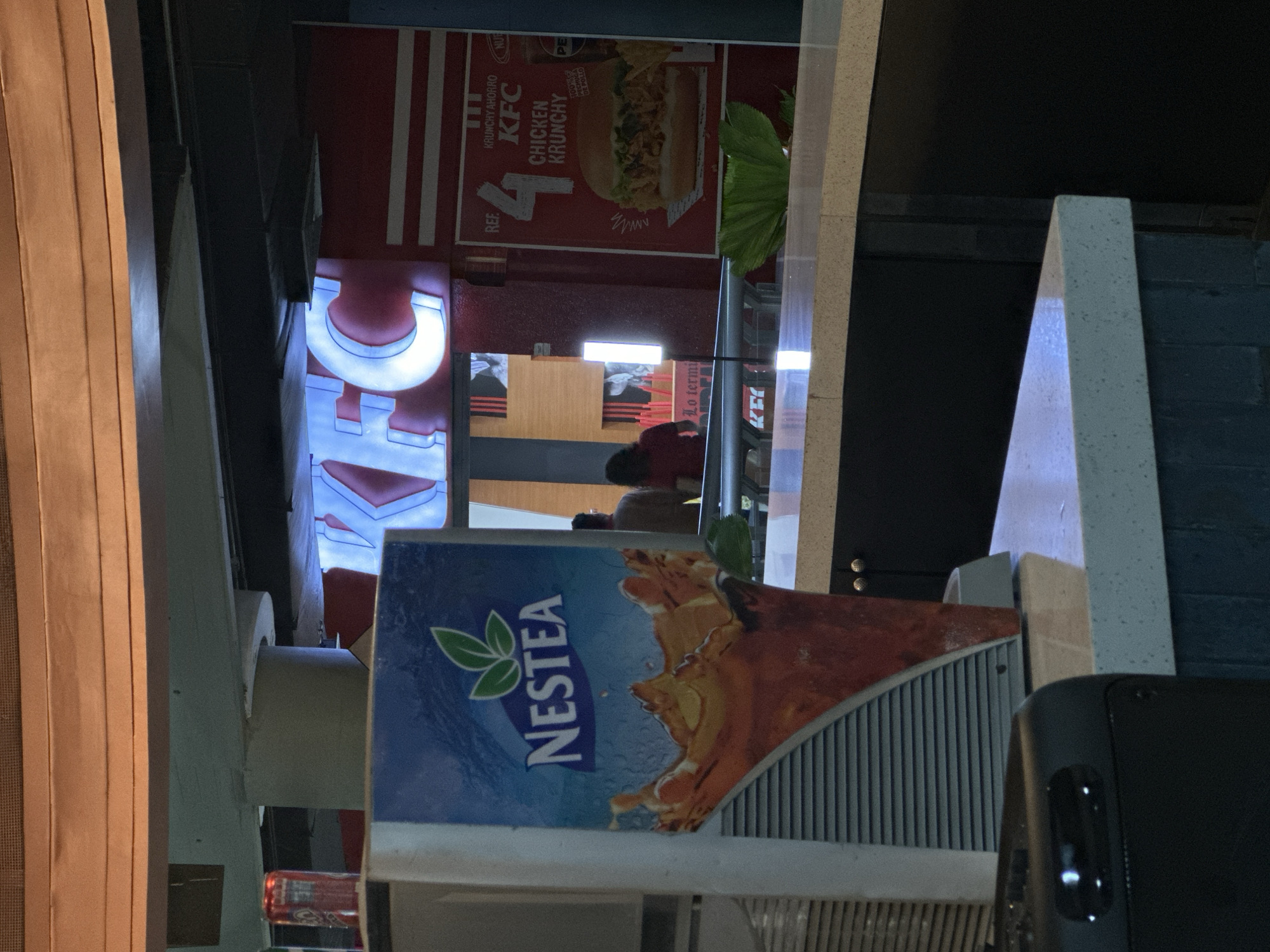 The image shows the exterior of a KFC restaurant. Prominent are the KFC logo, an advertisement for the "Chicken Krunchy," and a Nestea vending machine. There are people visible inside the restaurant. The location is most likely Caracas, Venezuela, as implied by the OCR text "Lo termi" and the presence of brands commonly found in that region.