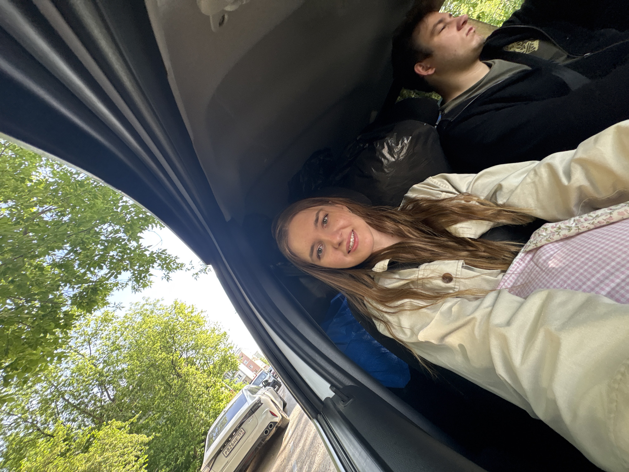 The interior of a car is depicted, with a young woman visible in the lower right portion of the frame. She is looking at the camera and smiling. She has long brown hair and is wearing a tan jacket over a black top. Part of a pink and white checkered blanket is also visible. An individual, presumed to be male, is lying down with his eyes closed in the upper right part of the frame. He has short dark hair and is wearing a black jacket over a dark green top. A black plastic bag is visible behind the woman.
Through the open car door, a tree with green leaves is visible. A white car with the license plate "P826HE 159" is parked on the side of the road. This license plate indicates the location of the photograph is Perm, Russia. A building can also be seen in the background.