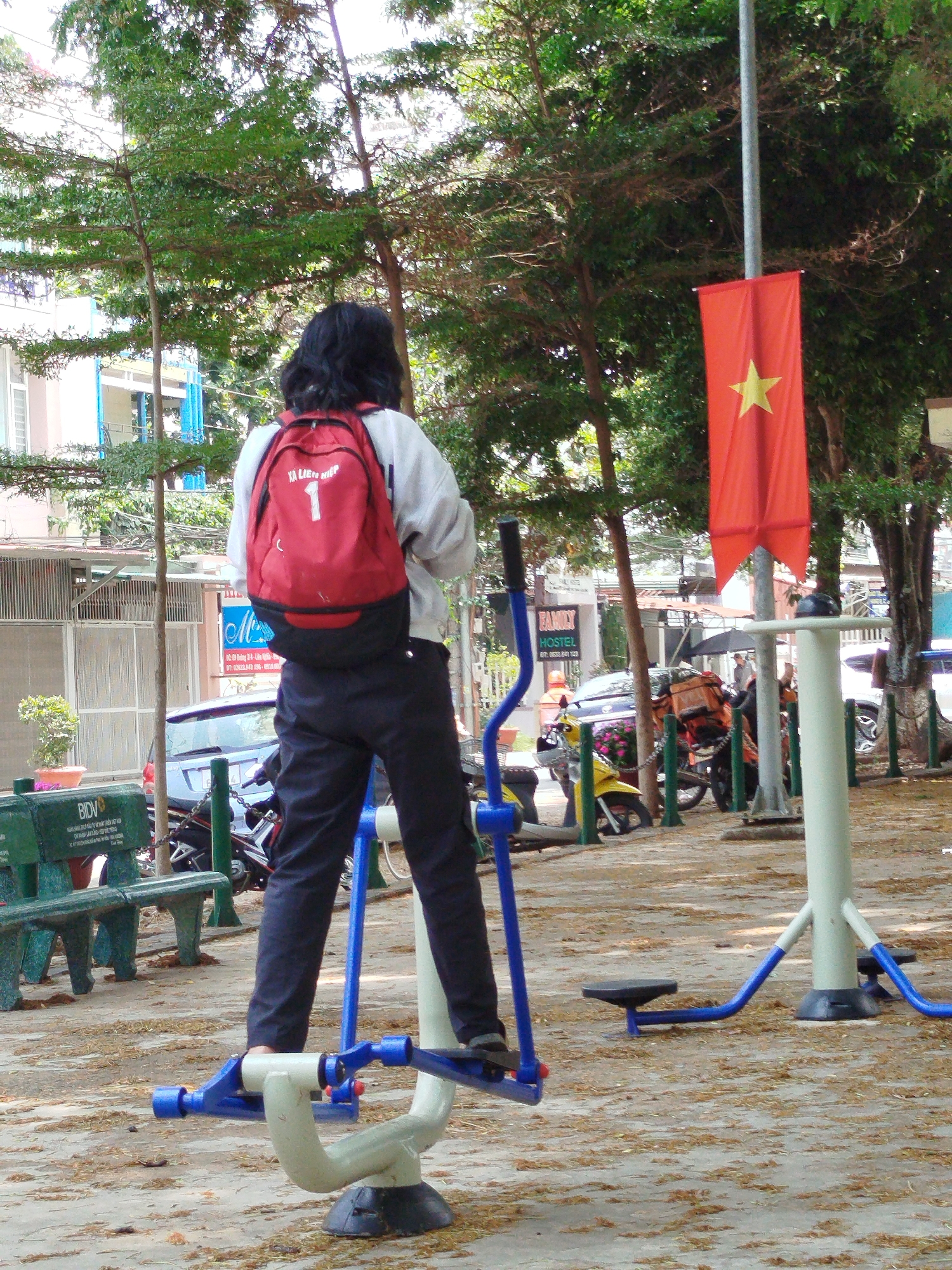 A person with a red backpack with the number 1 on it is using an outdoor exercise machine. The scene is set in a public space with trees and benches. In the background, there are parked vehicles, and a red flag with a yellow star (the flag of Vietnam) is prominently displayed on a pole. This place is located in Liên Nghĩa, Vietnam.