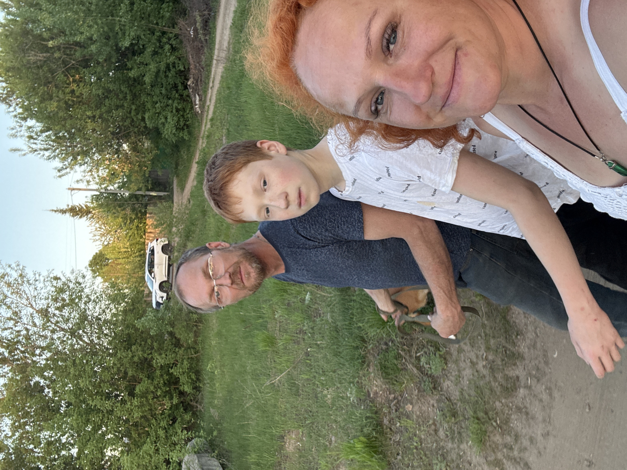 A selfie photograph shows a woman with red hair and a white top holding the camera, with a young boy with red hair standing behind her. A man with glasses and a blue shirt is slightly behind the boy, holding a dog leash. They appear to be outdoors in a grassy area with trees, near a dirt road. A white car is parked in the background. No specific details in the image indicate it is in Chaikovsky, Russia.