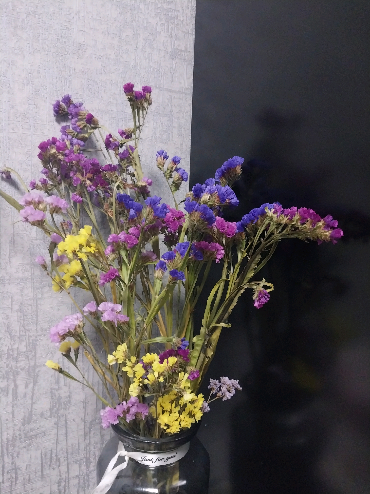 A bouquet of dried flowers, primarily in shades of purple, blue, and yellow, is arranged in a dark glass vase. The vase has a white ribbon tied around its neck with the text "Just for you" printed on it. The background is divided into two parts: on the left, a grey textured wall, and on the right, a solid black surface. The lighting suggests an indoor setting, likely a room. The image does not depict any specific location within the city of Dnipro, Ukraine. There are no people or actions visible in the scene.