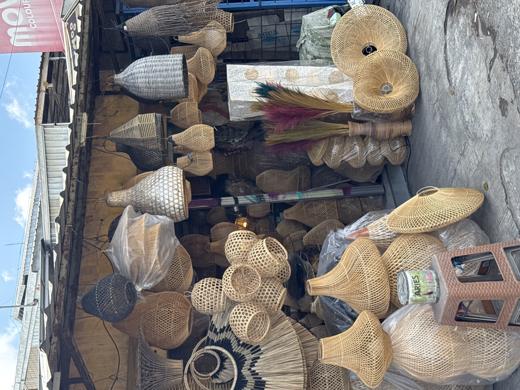 This is a medium-wide, eye-level shot of a street-side stall in North Kuta, Indonesia, selling a variety of woven rattan lampshades and decorative items. The stall appears to be open-air, with a corrugated metal roof supported by a rusted metal frame. The background consists of the stall's structure and some packaging materials.
In the foreground and midground, there is an abundance of rattan products. Hanging from the stall's roof are numerous lampshades of various shapes and sizes, predominantly in natural rattan tones, but also some in black and white. These range from large, bell-shaped ones to smaller, more intricate spherical designs. Some lampshades are covered in plastic for protection.
On the ground, more rattan items are displayed, including large, flat, circular lampshades and stacked lampshades of different designs. A small, brown plastic stool sits in the mid-ground, topped with a rusty, cylindrical metal container labeled "ARIES" with a faded graphic of a garden scene. To the right of the stool, there are decorative dried reeds with colorful tips (pink, green, and dark purple) arranged in two vases. Behind the reeds, a large, patterned white fabric is displayed, possibly a textile or a backdrop.
The ground is a concrete surface with visible cracks and water stains. The overall impression is of a bustling marketplace with a focus on artisanal crafts. There are no people visible in the image. The lighting suggests it is daytime with a bright sky, indicated by shadows and highlights.