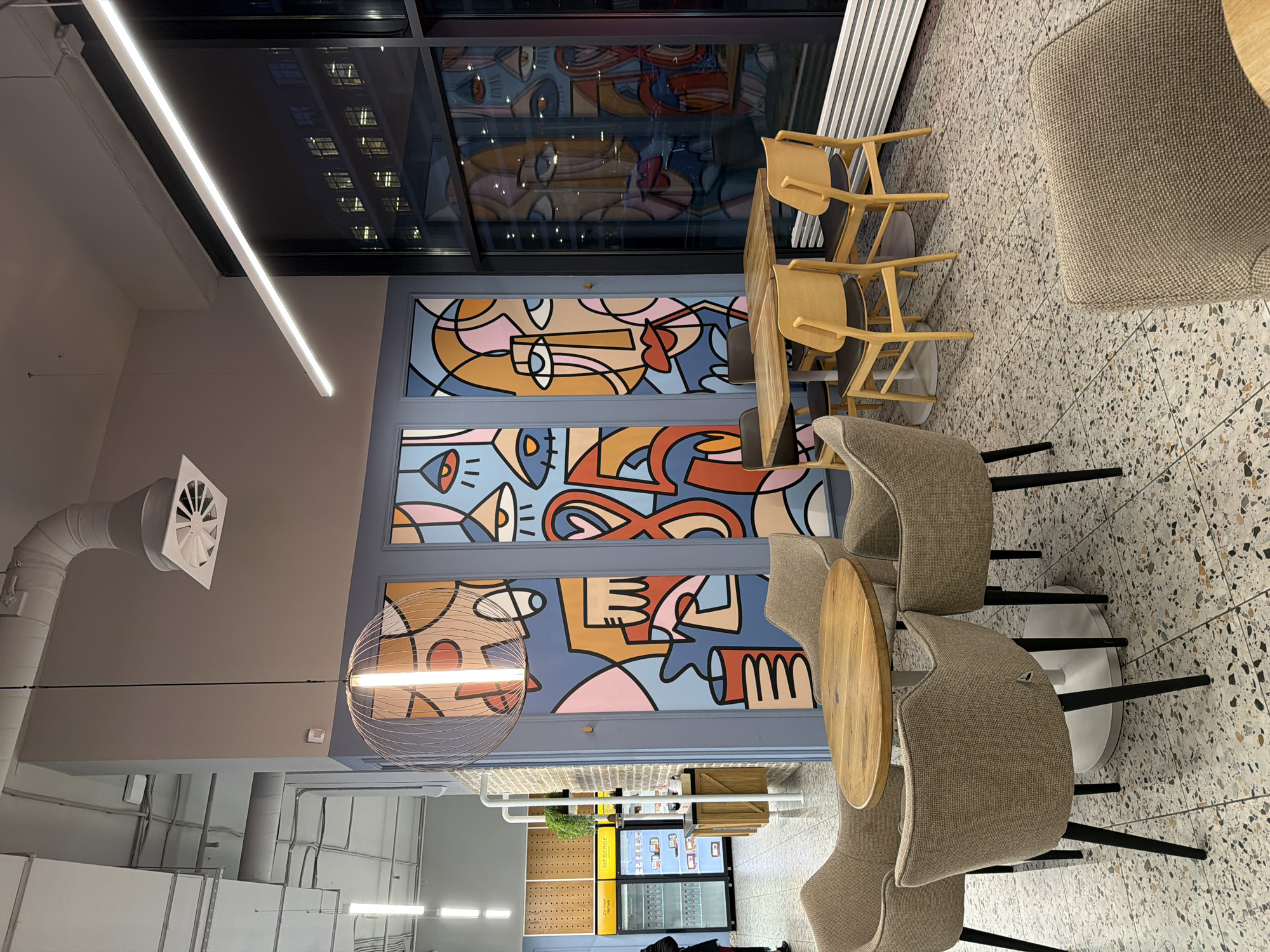 The image shows the interior of a cafe or restaurant in Moscow, Russia. There are tables with chairs set for dining, and a colorful mural on the wall depicting abstract faces or figures. A large window reflects the city lights and building exteriors. The decor is modern with exposed ductwork and decorative lighting fixtures. The floor is a patterned terrazzo. A menu board and other fixtures suggest it is a functioning eatery. There are no people immediately visible in the image.