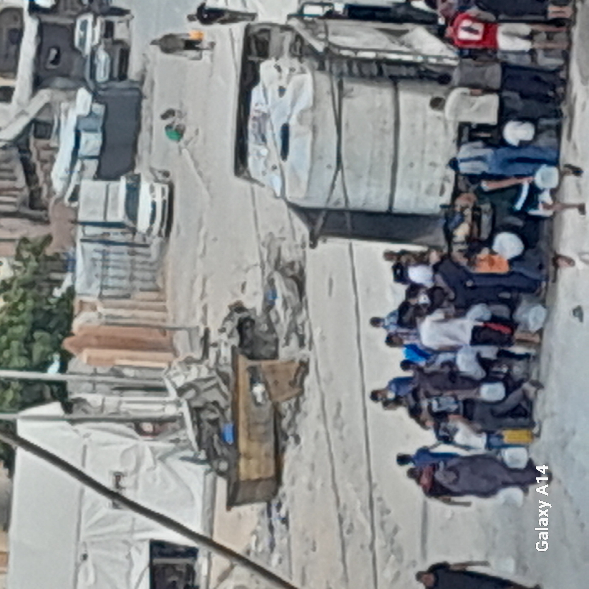 In an outdoor scene in an unknown location, possibly a city street or a makeshift camp, a group of people are gathered around a large truck with multiple water tanks. The ground appears to be dusty or sandy. Several individuals are holding white or clear plastic containers, suggesting they are collecting water. Some people are interacting with each other, while others seem to be waiting. In the background, there are tents, damaged structures, and vehicles, including a white car and a pickup truck. The overall impression is one of a community accessing essential resources.