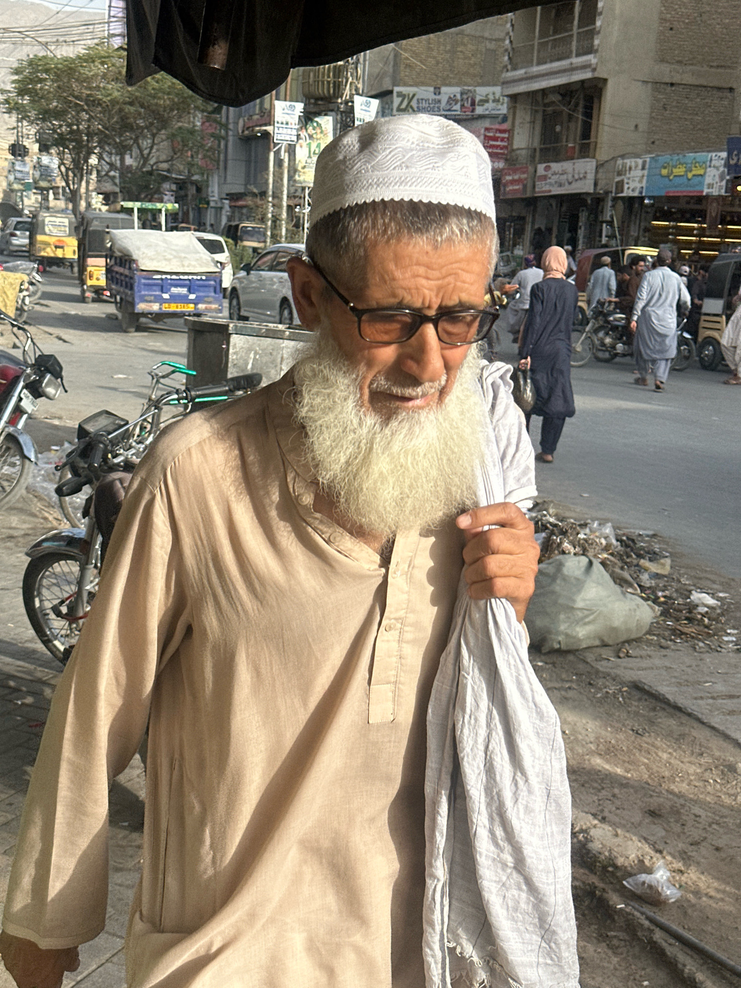 The image shows an elderly man with a long white beard, wearing a white prayer cap and glasses, standing on a street in Quetta City Tehsil, Pakistan. He is wearing a light brown tunic and holding a white cloth in his right hand. The background shows a busy street scene with various vehicles, including a blue truck, yellow auto-rickshaws, and motorcycles. Several other people are visible in the background, some walking and others on motorcycles. Buildings line the street, with shops and advertisements displayed. There is a pile of garbage on the right side of the image. The lighting suggests it is daytime, with sunlight casting shadows.