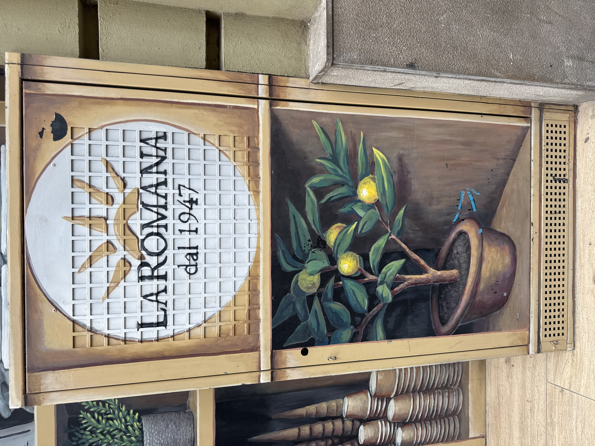 The image shows the exterior of a street cabinet in Valencia, Spain, decorated with a mural. The top section of the mural depicts the text "LAROMANA dal 1947" within a circular frame against a grid background, framed by a faux wood border. The bottom section features a painted image of a potted lemon tree with several yellow lemons and green leaves. To the left of the street cabinet, a portion of another cabinet is visible, painted to resemble stacked cups or cones. The street cabinet itself is made of metal and appears to be a utility box, with ventilation grates at the bottom. The surrounding environment includes a textured concrete wall to the right and a tiled floor at the bottom.