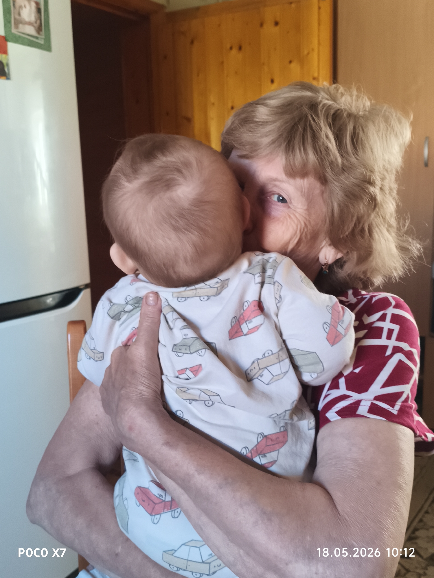 An elderly woman with short, curly blonde hair holds a baby wearing a white onesie with a car print design. The woman is wearing a red and white patterned short-sleeved shirt. She is looking at the camera, and her face is pressed against the baby's head, as if she is kissing the baby. The location appears to be inside a home, possibly a kitchen. In the background, there is a white refrigerator, wooden cabinetry, and a doorway. The bottom left corner of the image has text that reads "POCO X7," and the bottom right corner has a date and time stamp "18.05.2026 10:12." While the text could indicate the location is in the city of Chaikovsky, Russia, this information is not confirmed by the image itself.