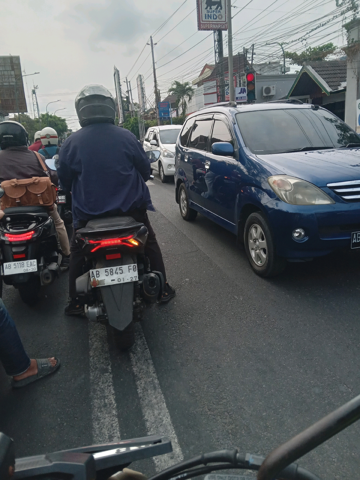 The image depicts a traffic scene in Yogyakarta, Indonesia, on a cloudy day. In the foreground, two motorcycles are stopped in traffic, one dark colored on the left and another dark colored with a gray rear fender on the right. The rider of the right motorcycle wears a dark blue jacket and a gray helmet. The rider of the left motorcycle wears a dark brown jacket and a black helmet. A brown saddlebag is attached to the rear of the left motorcycle. A blue minivan is in the right lane, to the right of the motorcycles. Behind the minivan, other vehicles and buildings line the street. A traffic light is visible, displaying a red light. A sign for "SUPER INDO SUPERMARKET" is visible above some buildings on the left side of the road. The road surface is asphalt with white lane markings. The general environment suggests an urban setting with moderate to heavy traffic.