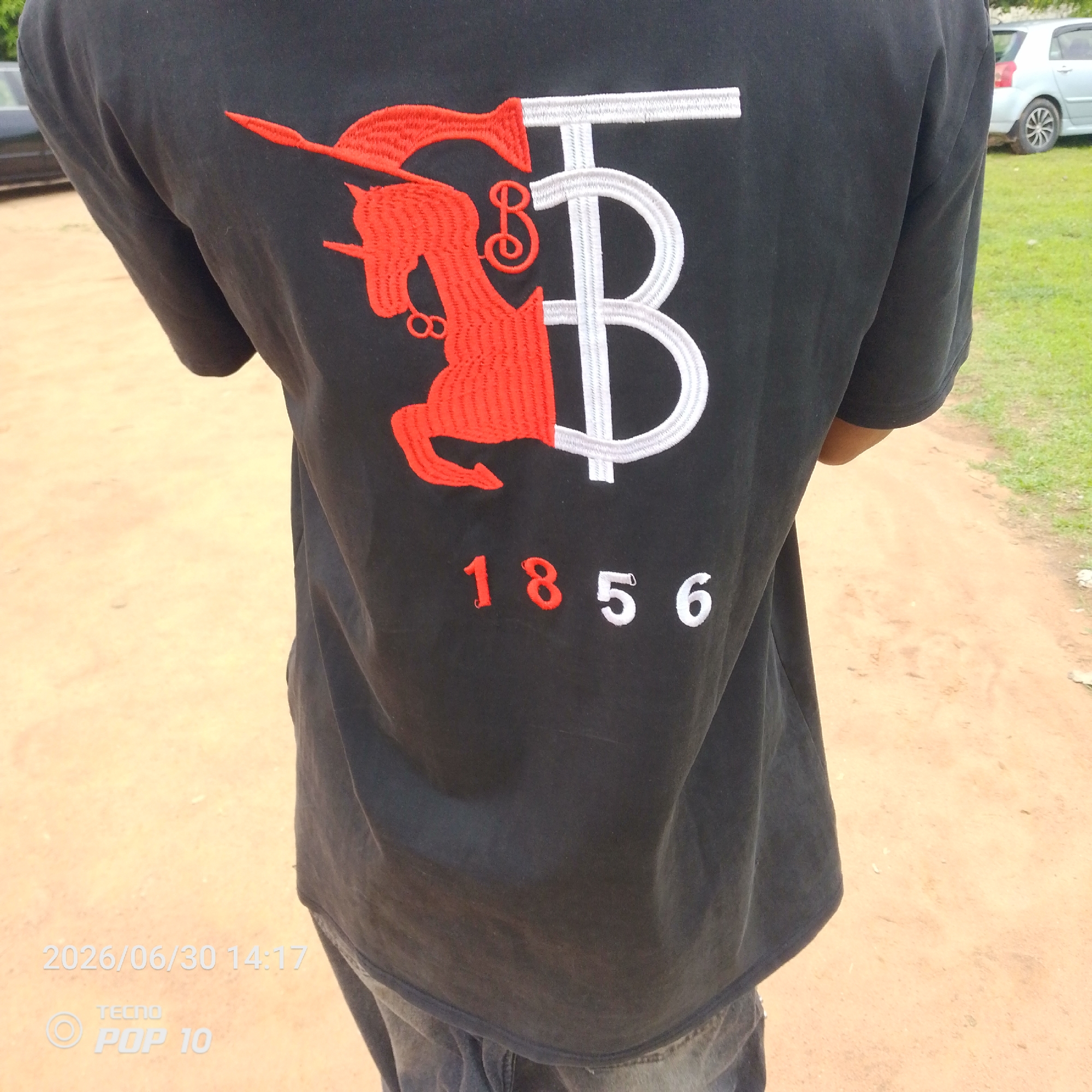 The image shows a person from behind, wearing a black t-shirt with an embroidered design. The design features a red unicorn on the left, with its head facing left and a horn pointing upwards. To the right of the unicorn is a large, stylized white "B." Below the unicorn and the "B" are embroidered numbers: "1856". The numbers "18" are red, and "56" are white.
The person is standing outdoors on a sandy path. To the right of the path, there is a grassy area with a silver car parked. The lighting suggests it is daytime. In the bottom left corner of the image, there is a timestamp and a phone model indicator: "2026/06/30 14:17" and "TECNO POP 10". The image appears to be taken in Jalingo, Nigeria.