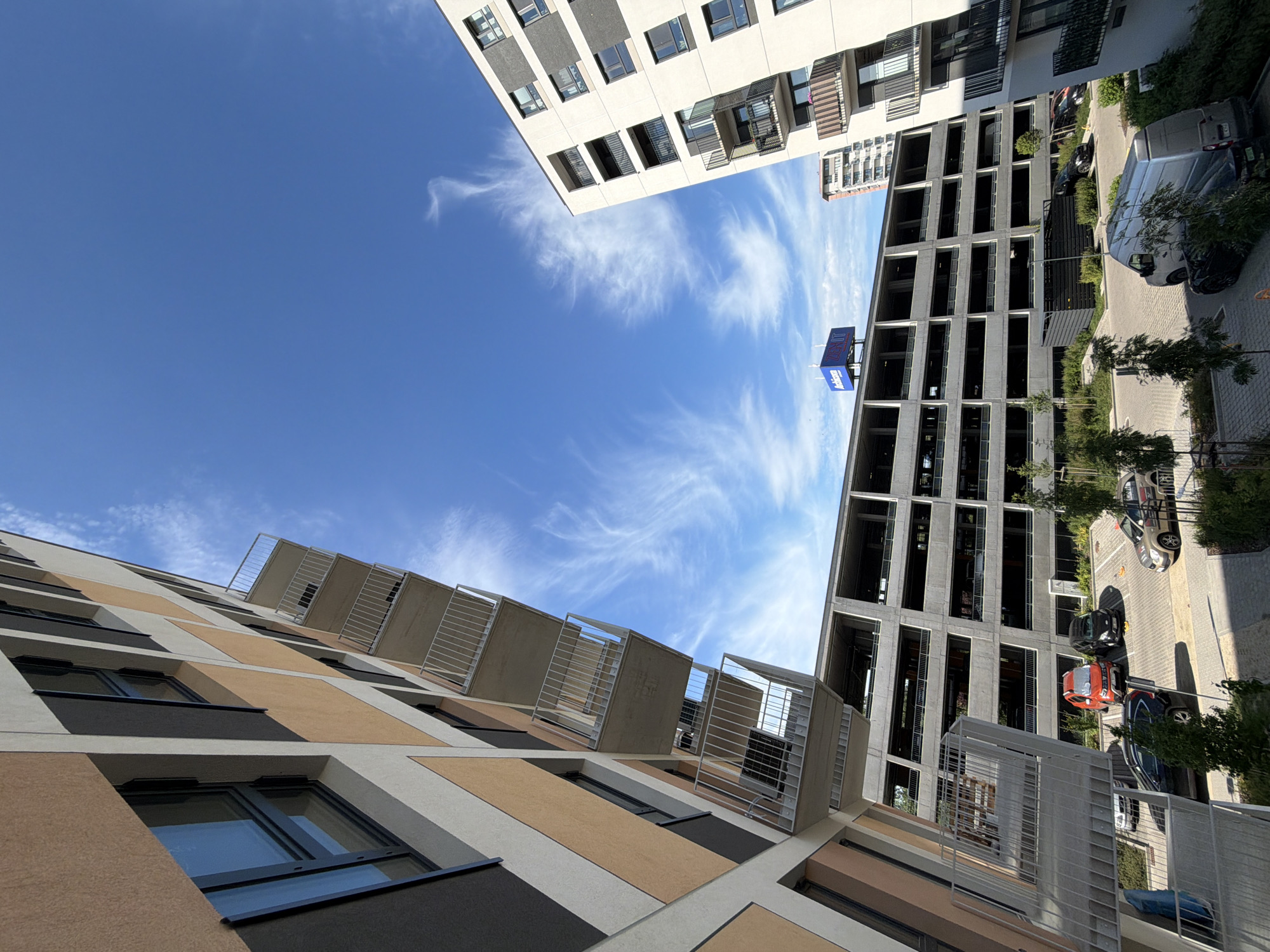 In a city scene, a modern apartment building with balconies and a multi-story parking garage dominate the frame. The apartment building, with its diagonal facade of tan, grey, and black panels, is seen from a low angle, with its balconies jutting out against a bright blue sky filled with wispy clouds. Adjacent to it is a large, grey concrete parking garage with open levels, its architectural lines sharp and angular.
Below the parking garage, several cars are parked on a paved lot. A red SUV, a black car, and a silver sedan are visible. A silver van is parked in the foreground, partially obscured by greenery. The environment is urban, with paved surfaces, sparse trees, and low bushes lining the parking area. In the background, another taller residential building is visible. A blue and white sign with text is mounted on top of the parking garage. The scene appears to be a residential or mixed-use development in Łódź, Poland, possibly part of a newer construction project. There are no people immediately visible in the scene.