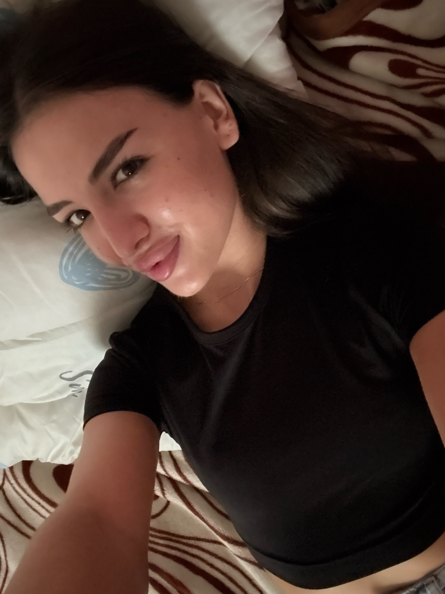 In Basarabeasca, Moldova, a young woman with dark hair is lying on a bed, looking towards the camera. She is wearing a black t-shirt. The bed is covered with a white pillow and a blanket with a brown and white striped pattern. The lighting is dim, suggesting an indoor setting, possibly at night or in a room with low light.