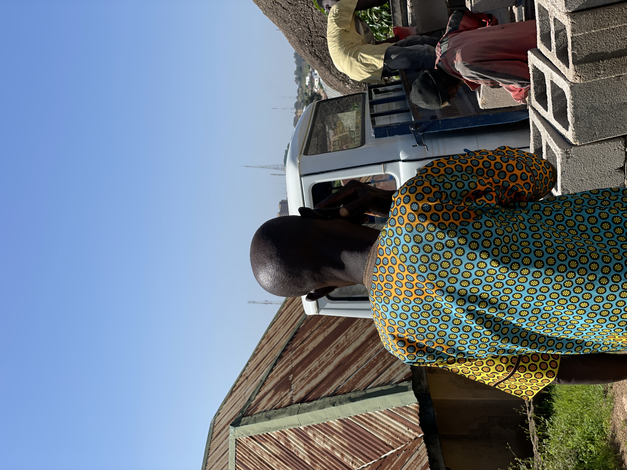 In the city of Jos, Nigeria, a man wearing a colorful patterned shirt is seen from behind, holding a phone to his ear. He stands on a dirt path next to a white pickup truck. In the background, two other men are visible, one in a yellow shirt bending down, and another in a red jacket with a black beanie, appearing to be working with concrete blocks. A corrugated metal roof of a building with green trim is to the left. The sky is clear and blue, and a rocky outcrop is on the right side of the image. The scene suggests an outdoor construction or work site.