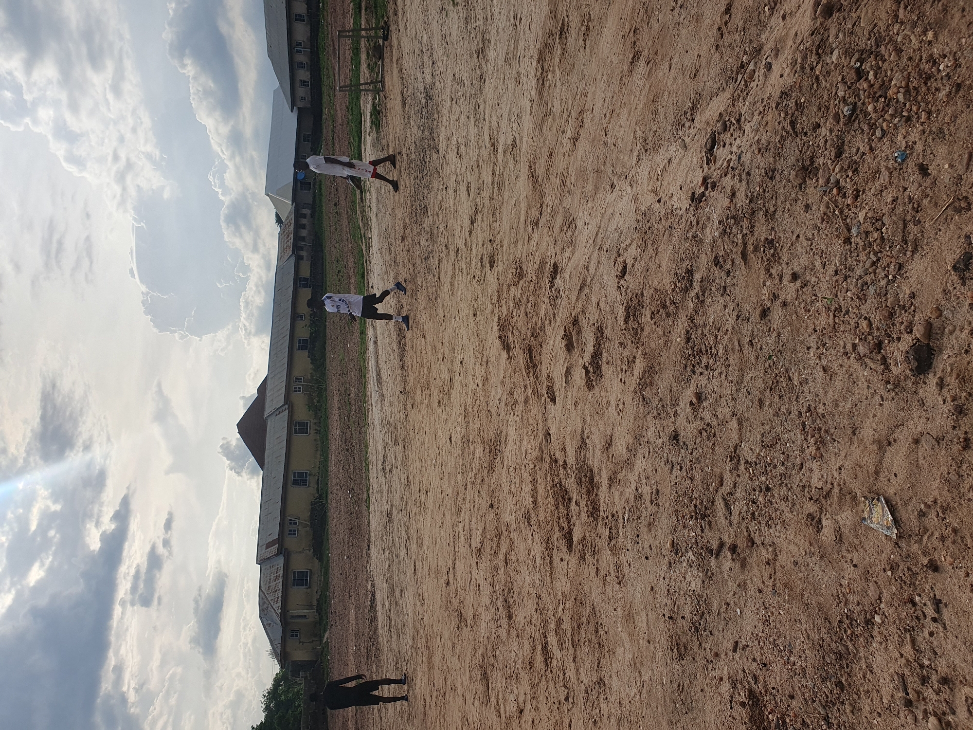 The image depicts an outdoor scene in Lokoja, Nigeria. Two individuals are seen on a sandy field, likely playing soccer. The ground is a mix of sand and pebbles. A building with a rusty tin roof runs along one side of the field. The sky overhead is filled with clouds. A soccer goalpost is visible in the background. There is garbage on the ground.