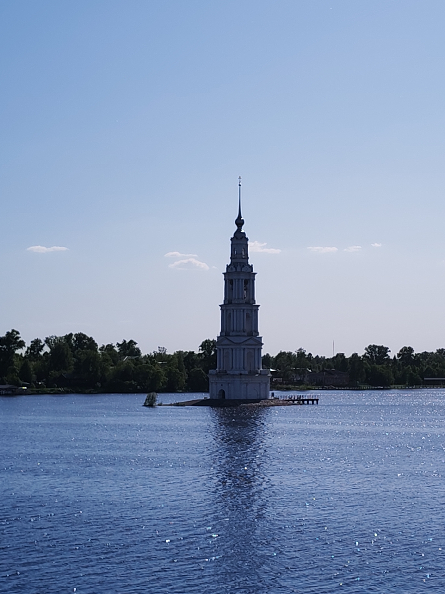The image depicts the flooded belfry of St. Nicholas Cathedral, a prominent landmark in Kalyazin, Russia. The tall, white, tiered structure with a spire and cross rises from the water. A small section of land and a simple wooden dock are visible near its base. The water is dark blue and shimmering with sunlight. A line of trees is visible on the horizon in the background. The sky is light blue with a few small white clouds. No people are visible.