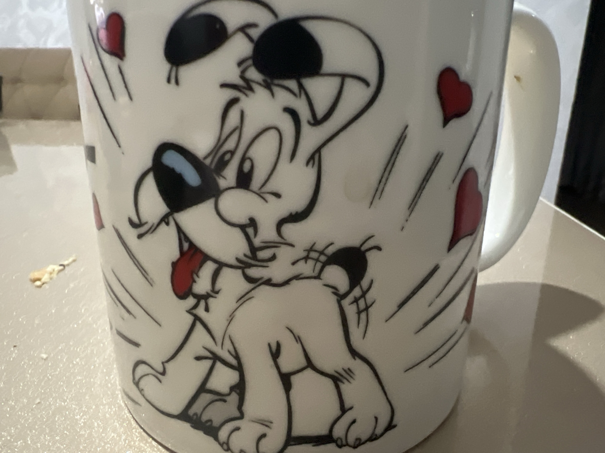 The image shows a white mug with a cartoon drawing of a dog on it. The dog is white with black accents and a red tongue. There are red hearts scattered around the dog. The mug is sitting on a light-colored surface. There are some crumbs on the surface to the left of the mug. The background is blurred. The mug is positioned on a table in an indoor setting.