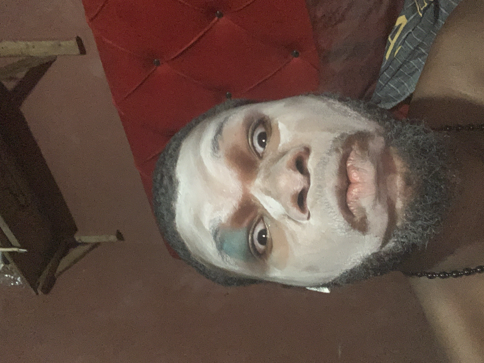 The image is a close-up, rotated portrait of an individual, likely male, with their face partially covered in a thick, white substance, resembling a face mask or theatrical makeup. A prominent dark, full beard frames the lower half of the face and cheeks. The person's eyes are open and look directly at the viewer with a neutral to slightly intense expression. A distinctive greenish-blue mark or shadow is visible near the inner corner of their left eye. The lips are naturally colored, appearing slightly parted.
In the background, a section of rich red, tufted upholstered furniture, possibly a headboard or chair back, occupies the upper right portion of the frame. A piece of grey fabric with yellow stripes, potentially part of clothing or a towel, is visible near the top right edge. The individual wears a dark beaded necklace, visible on the right side of their neck. Faint wooden structures are discernible in the background to the left, indicating an indoor setting with relatively dim or artificial lighting. No discernible text is present in the image. The scene takes place in Nkpor, Nigeria.