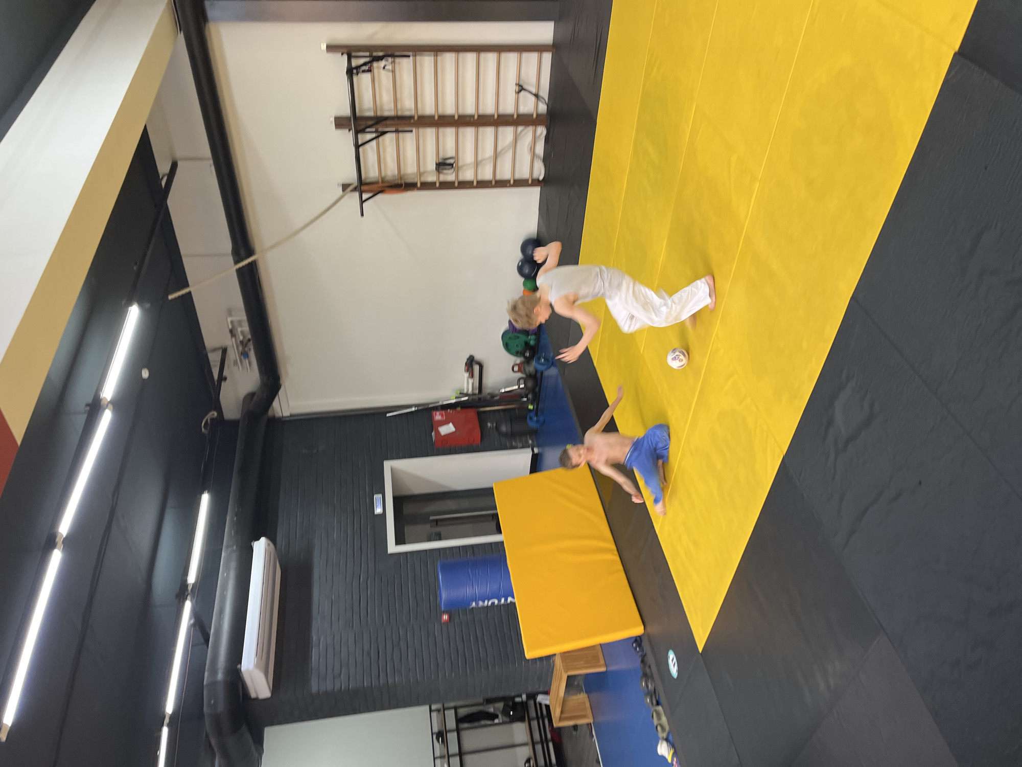 Two young boys are on a yellow mat in what appears to be a martial arts or gymnastics gym. One boy is seated on the mat wearing blue pants. The other boy is standing and appears to be kicking a soccer ball. The gym has black and gray mats on the floor, a black ceiling with fluorescent lights, and a white wall. There are gym equipment visible in the background, including a wooden wall ladder and various weights. This gym is located in the city of Krasnoyarsk, Russia.