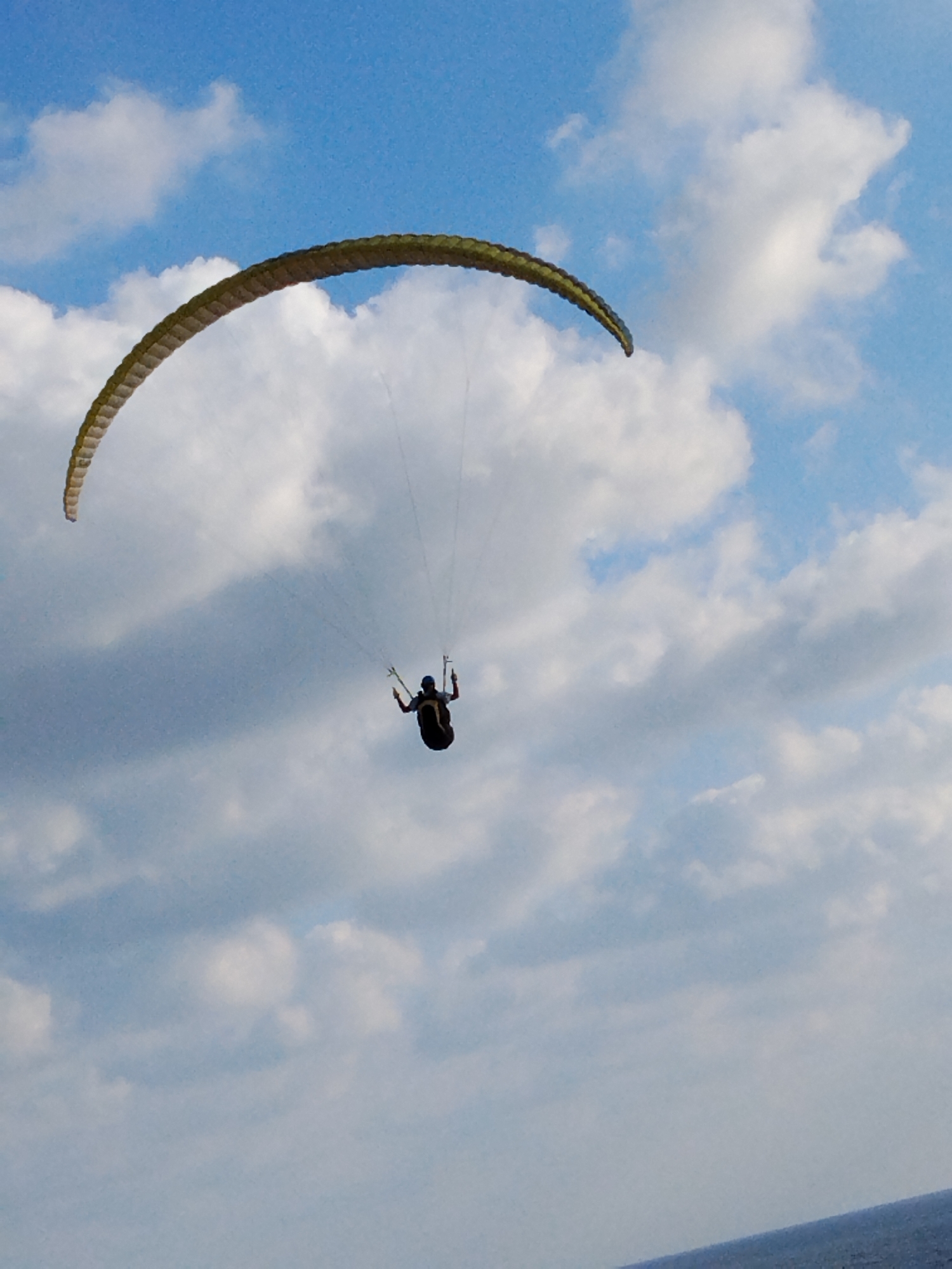 A paraglider is captured mid-flight against a backdrop of a blue sky dotted with white clouds. The paraglider, a person in a dark suit, is suspended beneath a large, curved parachute-like wing, which is predominantly light brown with dark green trim. The wing is positioned in the upper left quadrant of the image, extending towards the right. The paraglider is in the center of the frame, facing away from the viewer, with arms outstretched to control the glider. The scene appears to be over water, with a sliver of dark blue visible at the bottom edge of the image, likely representing the sea. The overall impression is one of freedom and open-air activity. This image was taken in Netanya, Israel.
