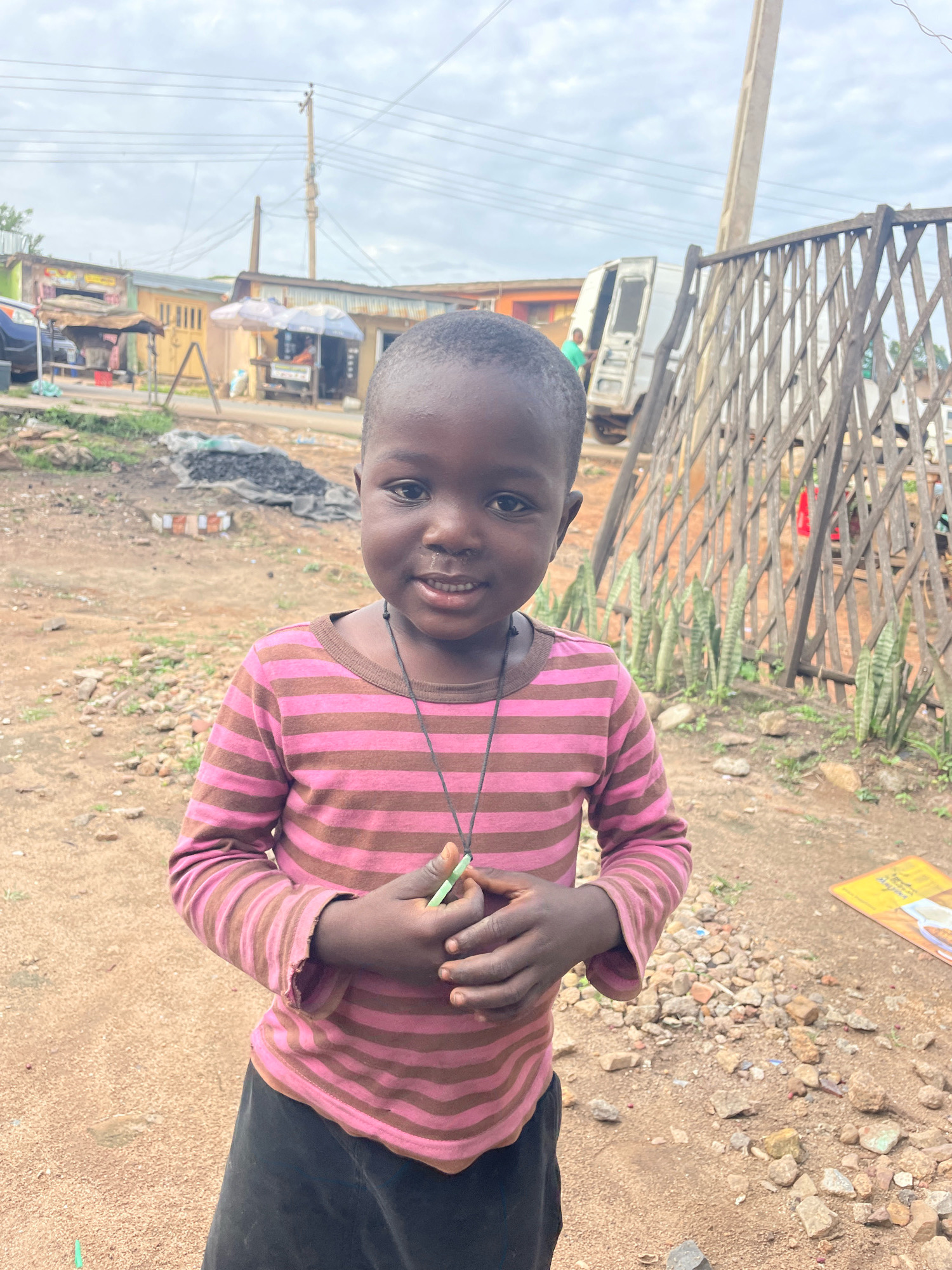 A young child, likely a girl, with dark skin and short black hair is standing outdoors, looking directly at the camera with a slight smile. She is wearing a long-sleeved pink and brown striped shirt and a dark skirt. A thin black string with a green pendant hangs around her neck. Her hands are clasped in front of her, and she appears to be holding a small green object.
The background suggests a semi-urban or rural environment. To the left, several market stalls with corrugated iron roofs are visible, some with awnings and merchandise displayed. A white van is parked to the right of the stalls, with its door open and a person in a green shirt visible inside. A tall utility pole with numerous wires stretches across the sky. In the foreground, the ground is a mix of dirt, small stones, and some darker debris. To the right of the child, a rustic wooden fence is visible, with several snake plants growing behind it. The overall lighting suggests an overcast day. The image appears to be taken in the city of Jos, Nigeria, given the context of the environment and the appearance of the child and surroundings.