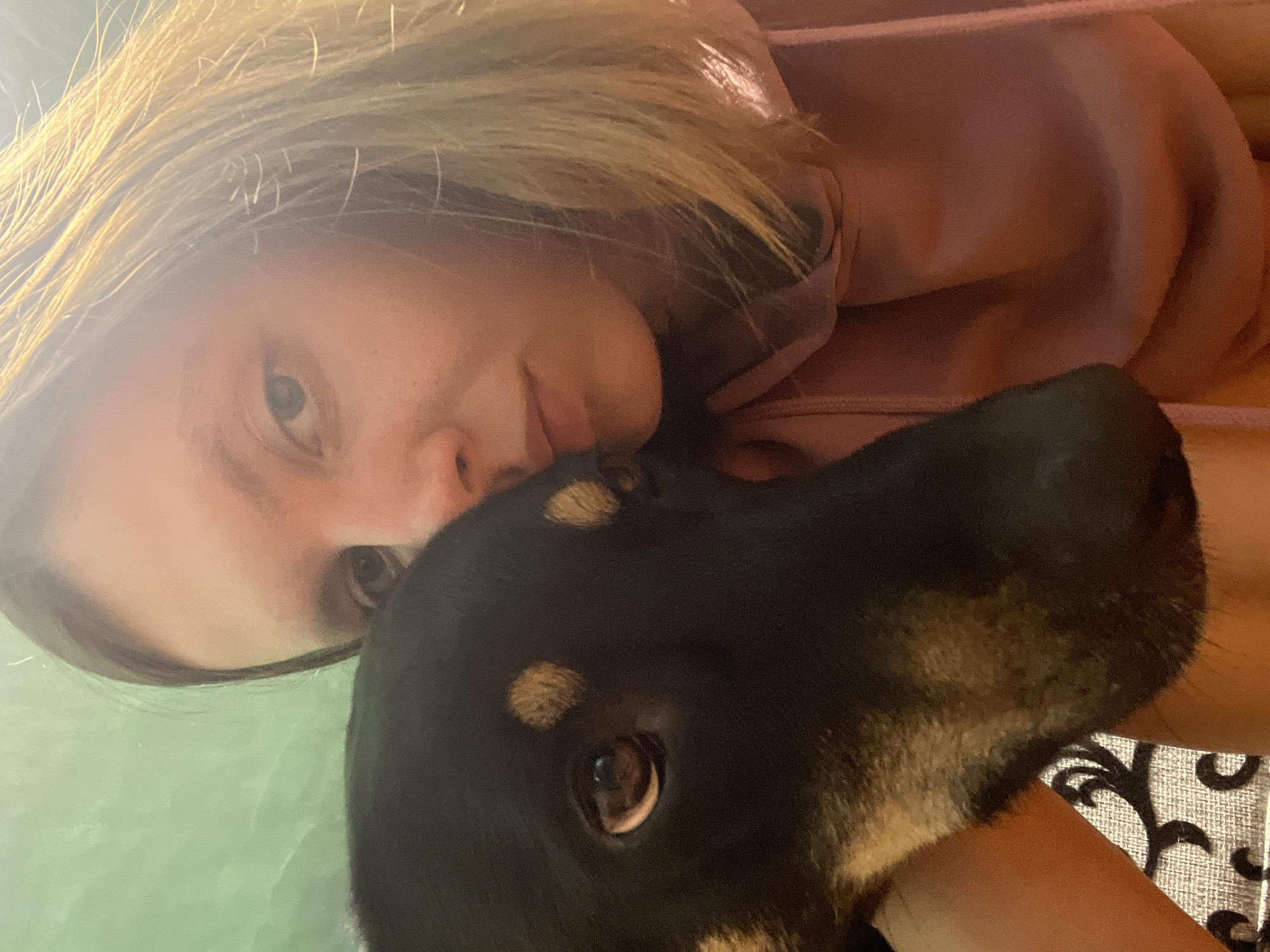 A close-up selfie shows a young woman with shoulder-length blonde hair and a pink hoodie, embracing a black dog. The dog's head, partially obscuring the woman's face, is turned towards the camera, revealing its brown eye and tan markings above its eyes and on its muzzle. The background consists of a textured green surface and the woman's patterned clothing. The lighting suggests an indoor setting, possibly daytime. The image was taken in Nizhny Tagil, Russia.