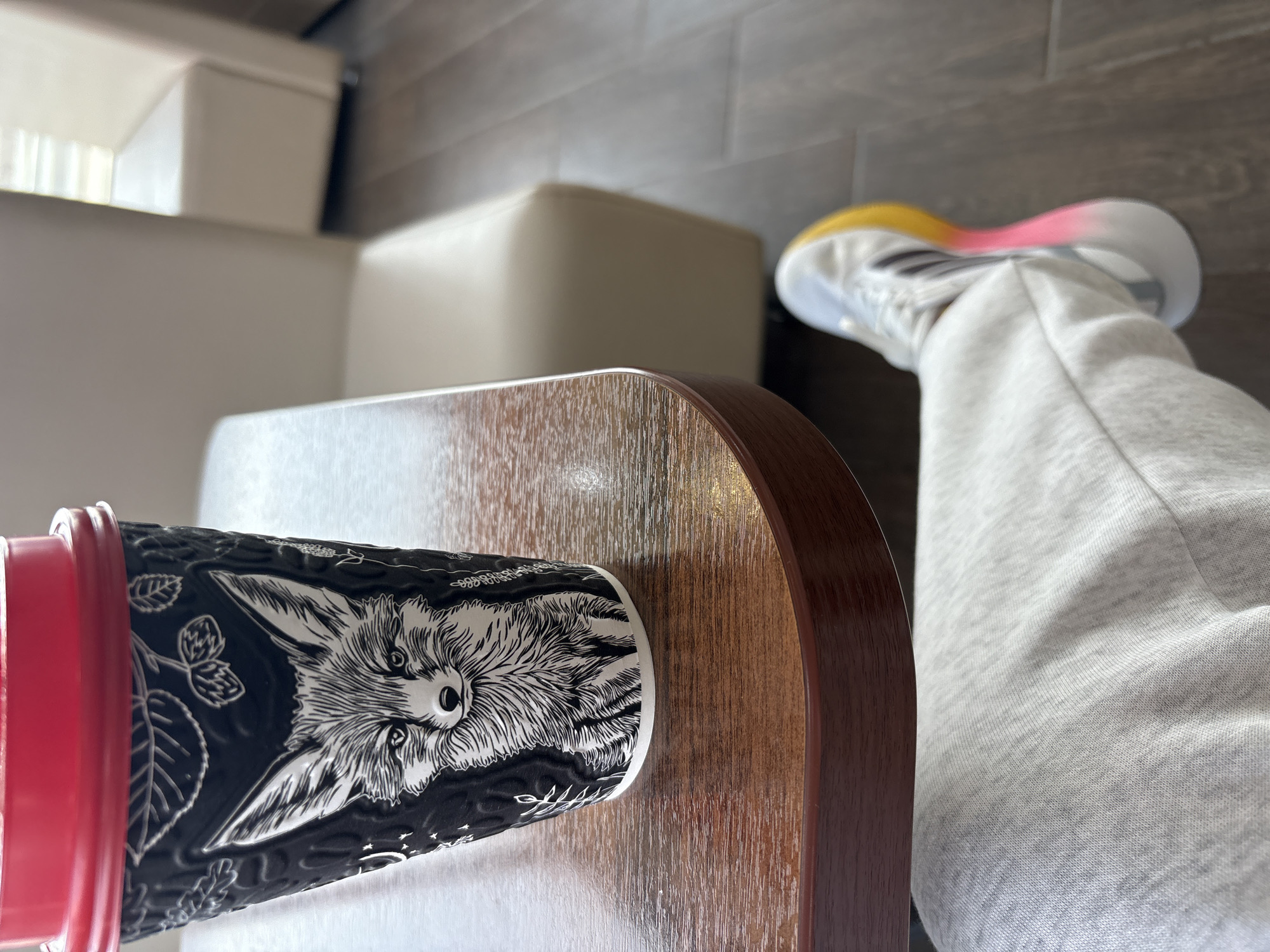 A cup with a black and white fox design rests on a wooden-toned table. A person wearing gray sweatpants and white athletic shoes with yellow and pink accents is positioned to the right of the table. White furniture is partially visible in the background. It is not possible to confirm a location in the city of Можга, Russia, based solely on the visual information provided.