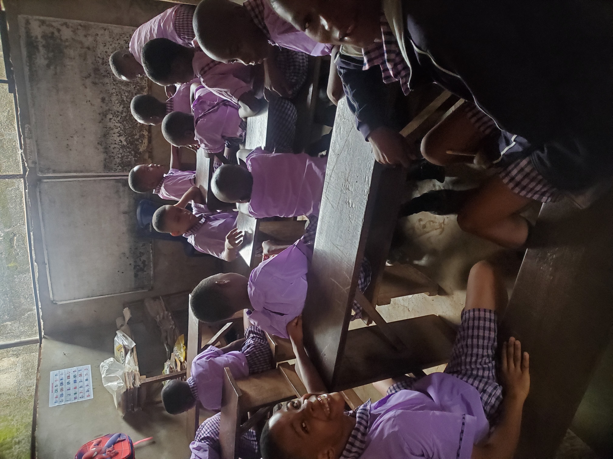 In Ukwu-Abwa, Nigeria, the image shows a classroom setting. A group of children, all wearing light purple shirts and checkered shorts or skirts, sit at wooden desks. The classroom appears somewhat dim and sparsely furnished. A poster is on the wall, and various items are stored in the corner. The students appear to be engaged in activities at their desks.