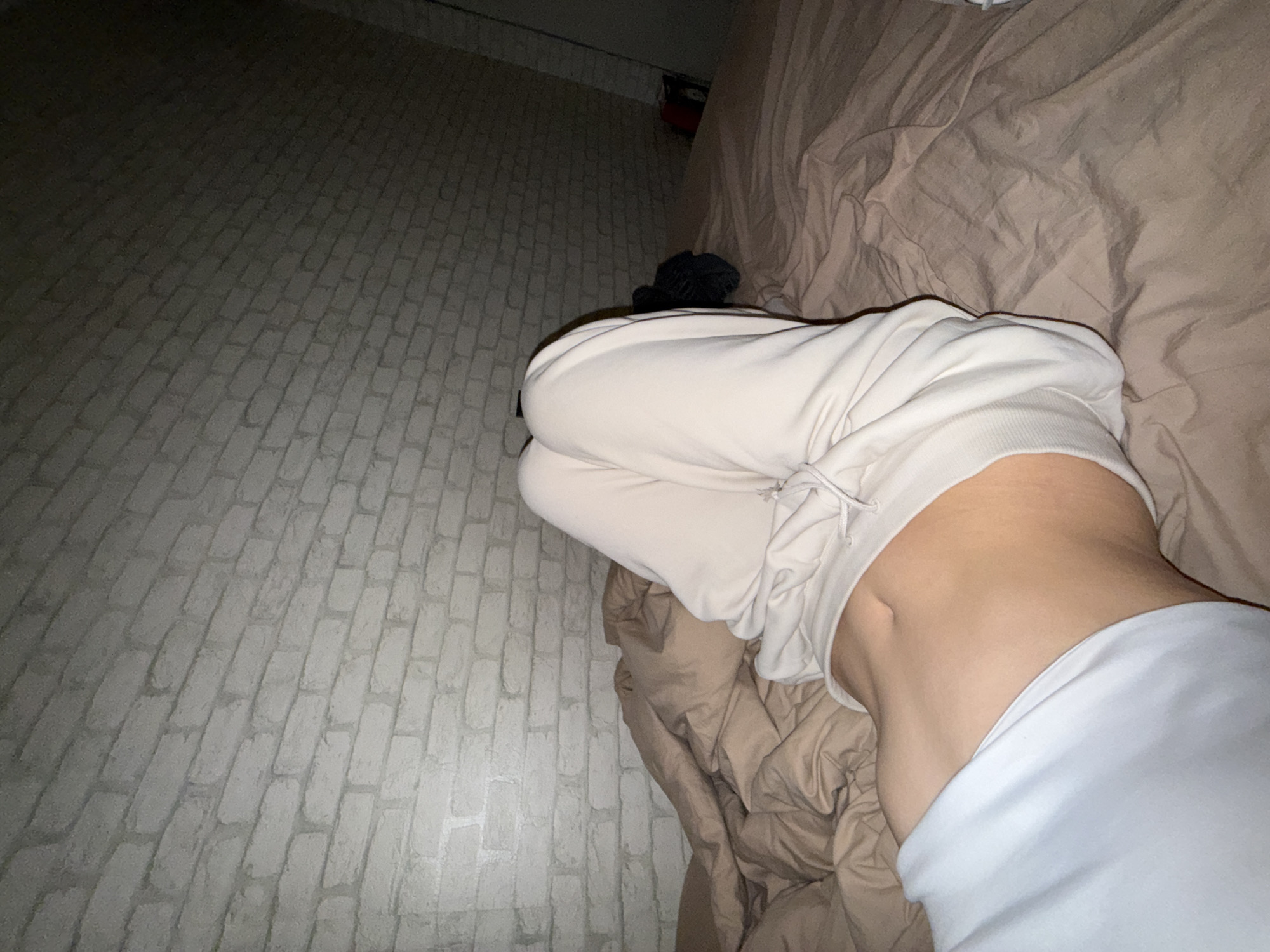 The image shows a person lying on a bed. The person is wearing light-colored sweatpants and a white sweatshirt. The bed has brown sheets and blankets. Behind the bed is a white brick wall. The lighting suggests it is nighttime or indoors with dim lighting. There are no discernible elements that indicate a specific location within the city of Sofia, Bulgaria.