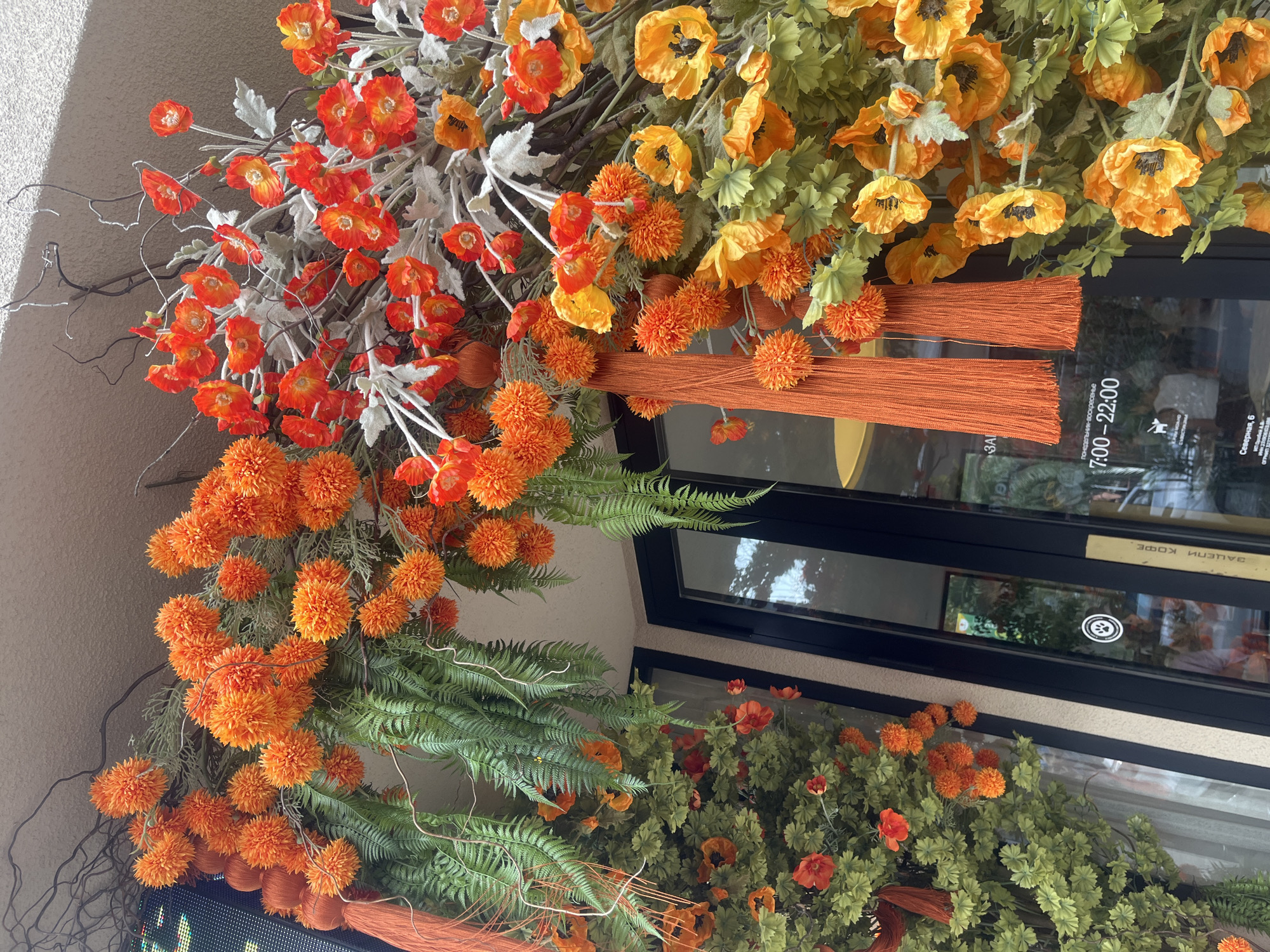 The image shows a storefront decorated with an abundance of artificial flowers in vibrant shades of orange and red, with green foliage. The floral arrangement is lush and cascading, featuring various types of blooms, including round, pom-pom-like flowers and delicate poppy-like flowers with dark centers. Several long, orange tassels hang from the arrangement, adding a festive touch.
The storefront itself is characterized by a large black-framed glass door, with white stripes on the exterior wall. A sign on the door displays operating hours in Russian: "ПОНЕДЕЛЬНИК-ВОСКРЕСЕНЬЕ 7:00–22:00". Another smaller sign with a paw print and text in Russian is visible below the hours. A reflection of the interior of the establishment, which appears to be a cafe or shop, is visible in the glass.
The location is identified as Sochi, Russia, at the address Severnaya, 6. The building exterior is a light-colored stucco. The overall impression is one of a welcoming and visually appealing entrance to a commercial establishment.
