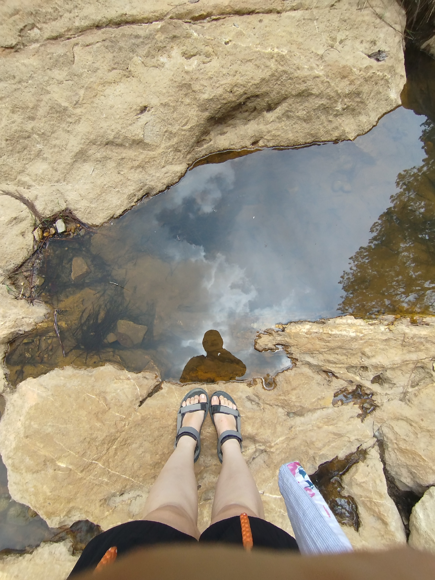 A person stands on a light brown, rocky surface, likely near or at the edge of a natural pool of water. The water reflects clouds and trees, with a dark silhouette of the person visible near the bottom of the reflection. The person is wearing grey sandals and dark shorts. The location of the image is Păng Tiêng 1, Vietnam. Additional objects include a striped object next to the person's right foot, sticks and leaves around the edge of the pool, and the surrounding rock formations.