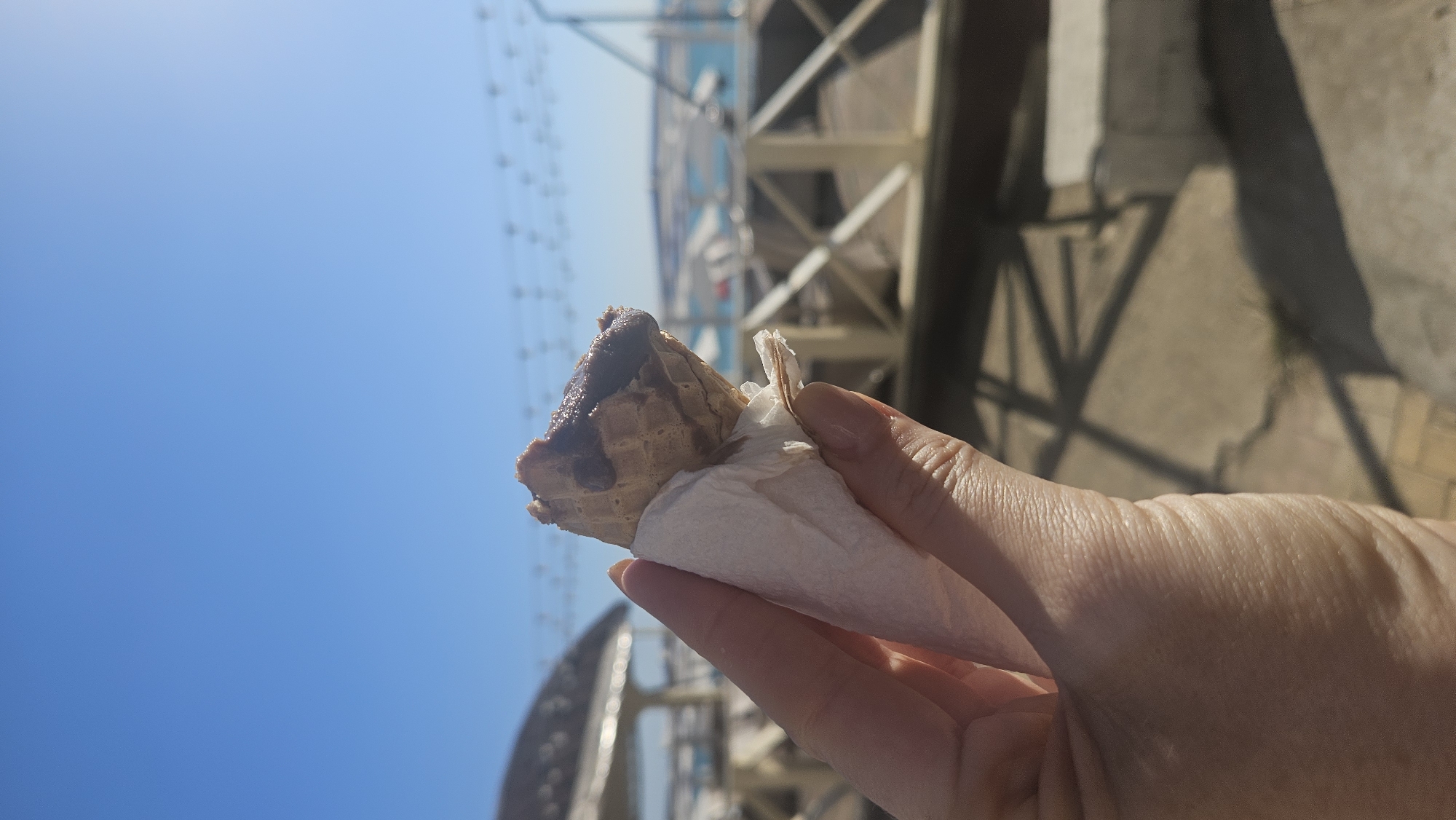 A hand holds a partially eaten chocolate ice cream cone wrapped in a white napkin against a clear blue sky. The cone is a wafer cone with dark chocolate drizzled on top. In the background, there is a fence, a gazebo or similar structure, and a body of water, possibly the sea. The ground appears to be paved. The scene is lit by bright sunlight.