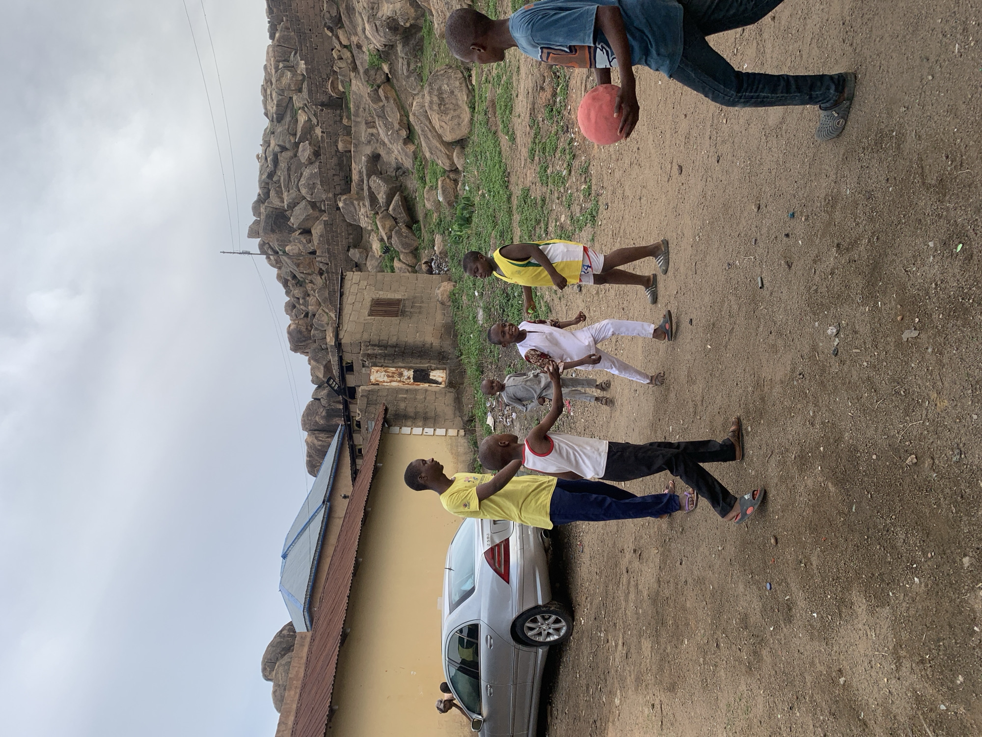Several boys are playing with a ball on a dirt lot, in what appears to be a residential area near Jos, Nigeria.
People:
- A group of young boys, some wearing shorts and T-shirts, others in more formal attire like a white suit.
- The boy at the top is holding a red ball.
Subjects:
- A light-colored car is parked next to a building.
- Rocky hills in the background.
Objects:
- A red ball, various types of clothing, and buildings.
Actions:
- The boys are playing, potentially passing the ball.
- Individuals are standing and moving.
Interactions:
- Boys are likely engaging in a game together, passing the ball.
Environment:
- A dirt lot serves as the playing field.
- Buildings, possibly houses, are located nearby.
- Rocky hills are visible in the distance.
- The image suggests an outdoor, somewhat rural setting within the city of Jos.