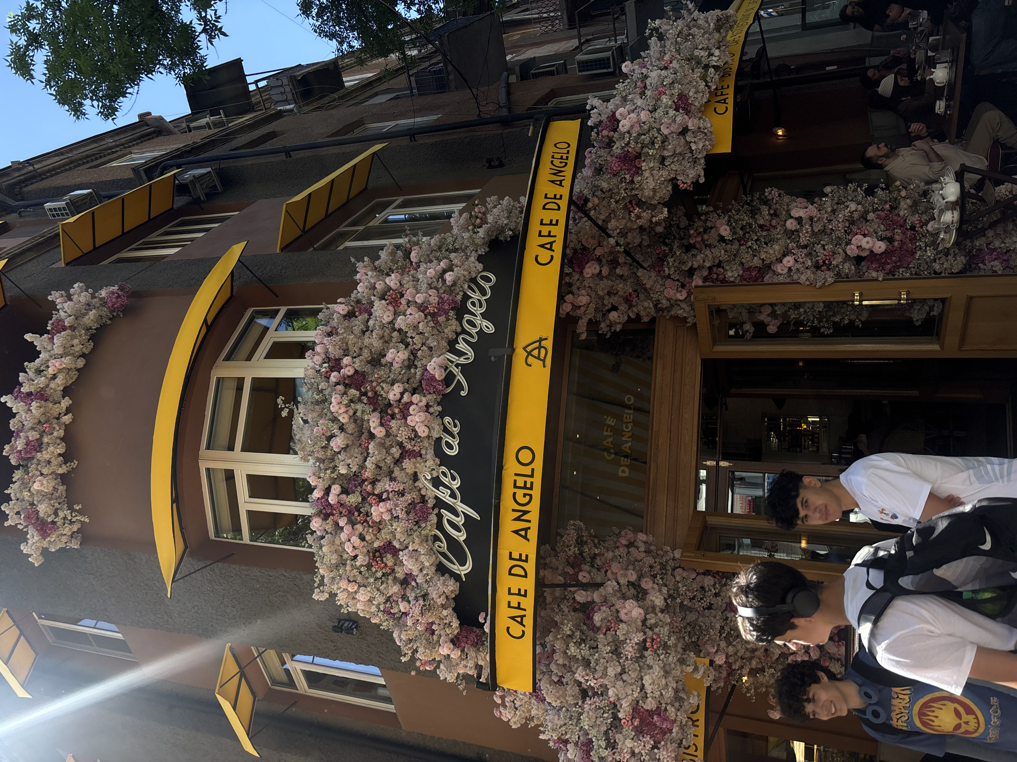 The image depicts the exterior of "Cafe de Angelo" in Yerevan, Armenia. The cafe's facade is adorned with abundant floral decorations, primarily pink and white blossoms. A yellow and black sign bearing the cafe's name hangs prominently. The architecture of the building includes windows with yellow awnings. In front of the cafe, three young people are walking, one wearing headphones and another with a backpack. The cafe's interior can be partially seen through the glass door, where other people are seated. The overall environment suggests an urban setting, with trees visible at the top.