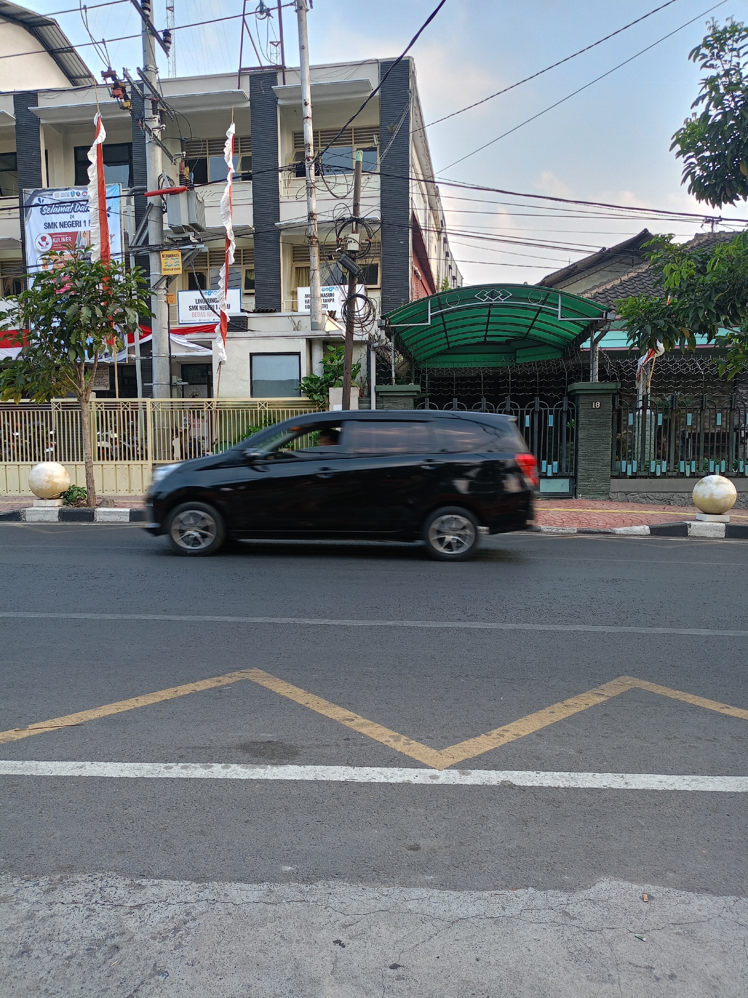 A black car is captured in motion on an asphalt road in front of a building. The building appears to be a school, with banners displaying "SELAMAT DATANG DI SMK NEGERI 1 BATU" (Welcome to SMK Negeri 1 Batu), indicating the location as Batu, Indonesia. The car is a minivan, with its wheels blurred due to movement. The road has yellow markings and a white line. On the sidewalk, a pale yellow spherical object stands next to a fence. The building features multiple stories with windows and a black facade. Electrical wires crisscross overhead, and trees with green foliage are visible on the right side of the frame. The sky is partially cloudy, suggesting it is daytime.