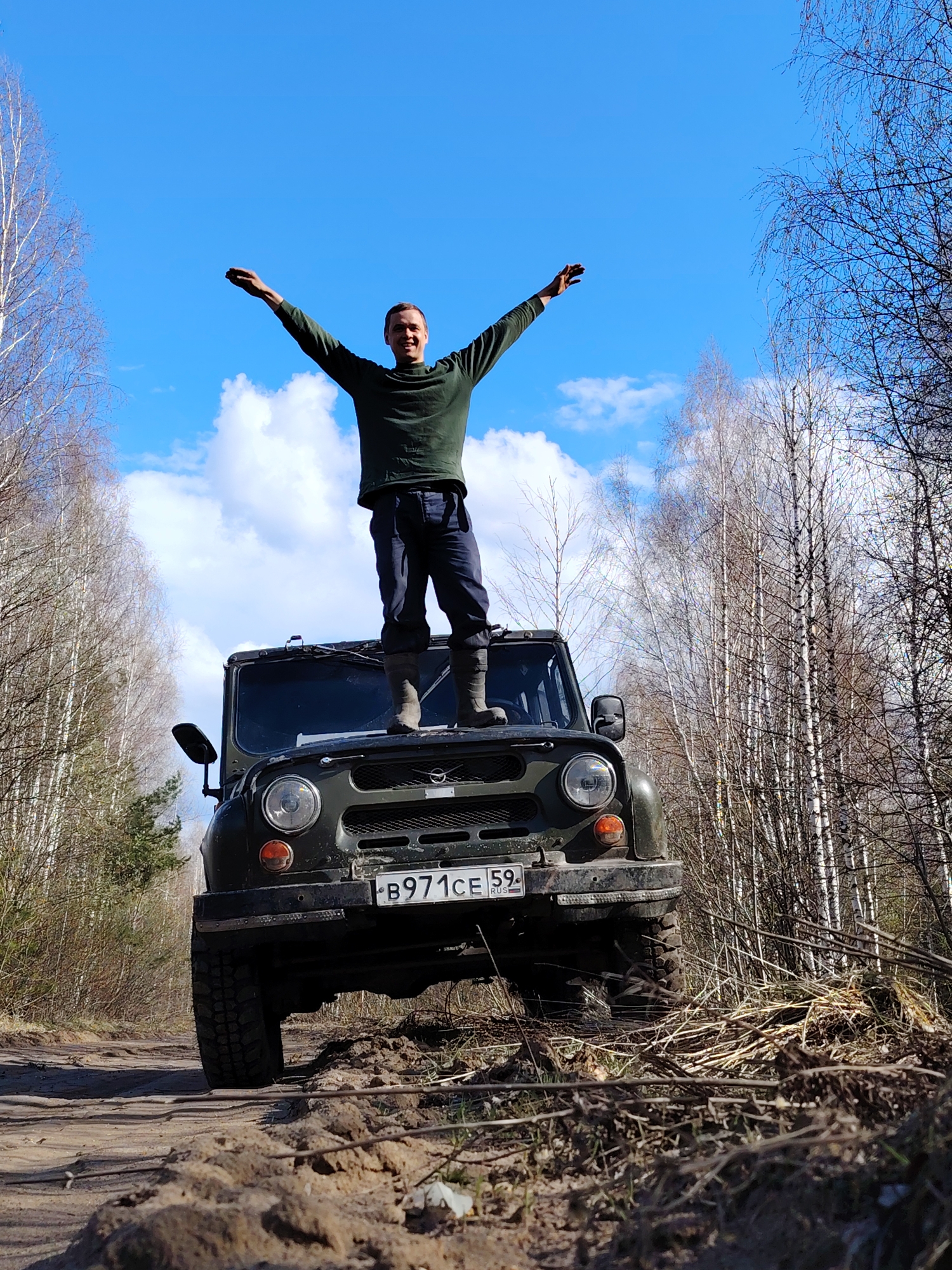 A man stands joyfully on the hood of a dark green off-road vehicle, his arms spread wide in a triumphant or celebratory gesture, with a wide smile on his face. He is wearing a dark green long-sleeved shirt, dark trousers, and sturdy boots.
The vehicle, which appears to be a UAZ model, is positioned on a dirt track in what looks like a woodland or forest setting. Its large tires are visible, suggesting its capability for rugged terrain, and the front bumper and grille are visible beneath the man's feet. A Russian license plate, "B971CE 59 RUS," is clearly legible on the front of the vehicle.
The surroundings are characterized by numerous bare deciduous trees, primarily birches with their distinctive white bark, indicating either early spring or late autumn. The sky is bright blue with scattered white clouds, suggesting a sunny and clear day. The ground beneath the vehicle is a rough dirt track with ruts and loose earth, typical of an off-road path. The scene conveys a sense of freedom, adventure, and enjoyment of the outdoors.