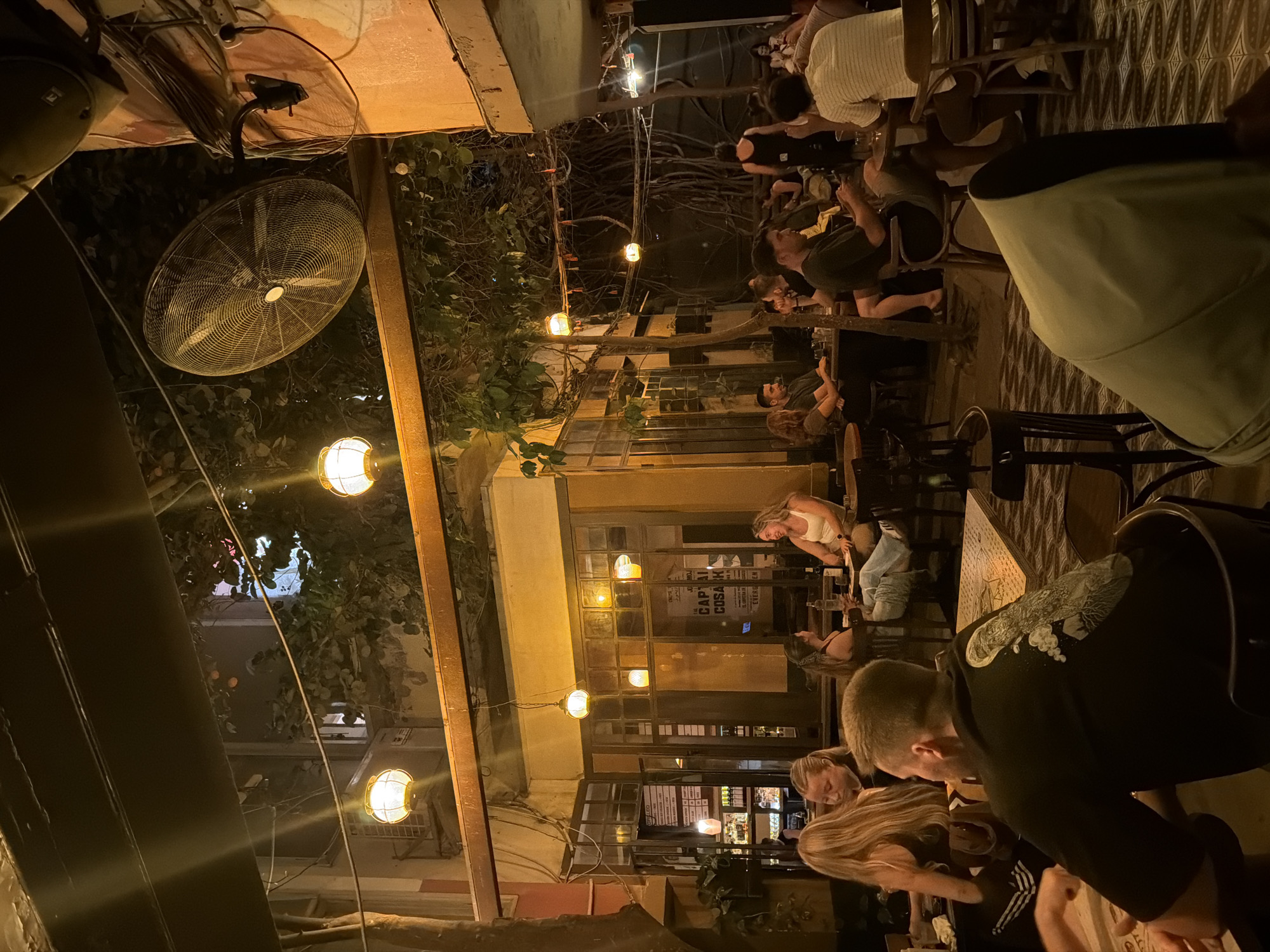 The image captures a lively outdoor or semi-outdoor bar or restaurant in Tel-Aviv, Israel, illuminated by artificial lighting at nighttime. Numerous individuals, predominantly young adults, are seated at tables, engaging in conversation, dining, and drinking in a casual, social atmosphere.
The setting features warm, spherical string lights hanging above and along the walls, casting a soft, inviting glow. A large, wall-mounted industrial-style fan is prominent on the left, suggesting a warm evening. Lush green foliage from trees or bushes is visible throughout the scene, intertwining with the light fixtures and architectural elements. Exposed electrical wiring and structural components are also discernible overhead.
The building facade in the background includes multiple windows. Through one window, a sign is clearly visible with text reading "GAP CUBA" and "CAPTAIN COSACK." Patrons are seen both inside the establishment through the windows and seated outside at wooden tables with various chair styles. The flooring in parts of the visible area features a distinctive patterned tile design. People are dressed casually, and many appear relaxed and engaged in their social interactions.