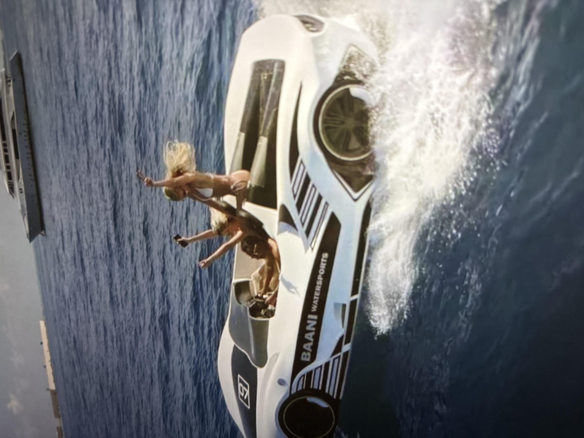 An unusual boat shaped like a sports car races across a body of water, kicking up a large spray. Two women are riding in the boat. The woman in the front, wearing a white bikini, has her arms raised in the air. The woman behind her, also in a white bikini, is holding her arms up and appears to be holding a phone. The boat is white with black and silver racing stripes and the words "BAANI WATERSPORTS" are printed on the side. In the background, a large white yacht is visible. The sky is blue with some white clouds. The scene is set on a sunny day. This image does not depict the city of Pori, Finland.