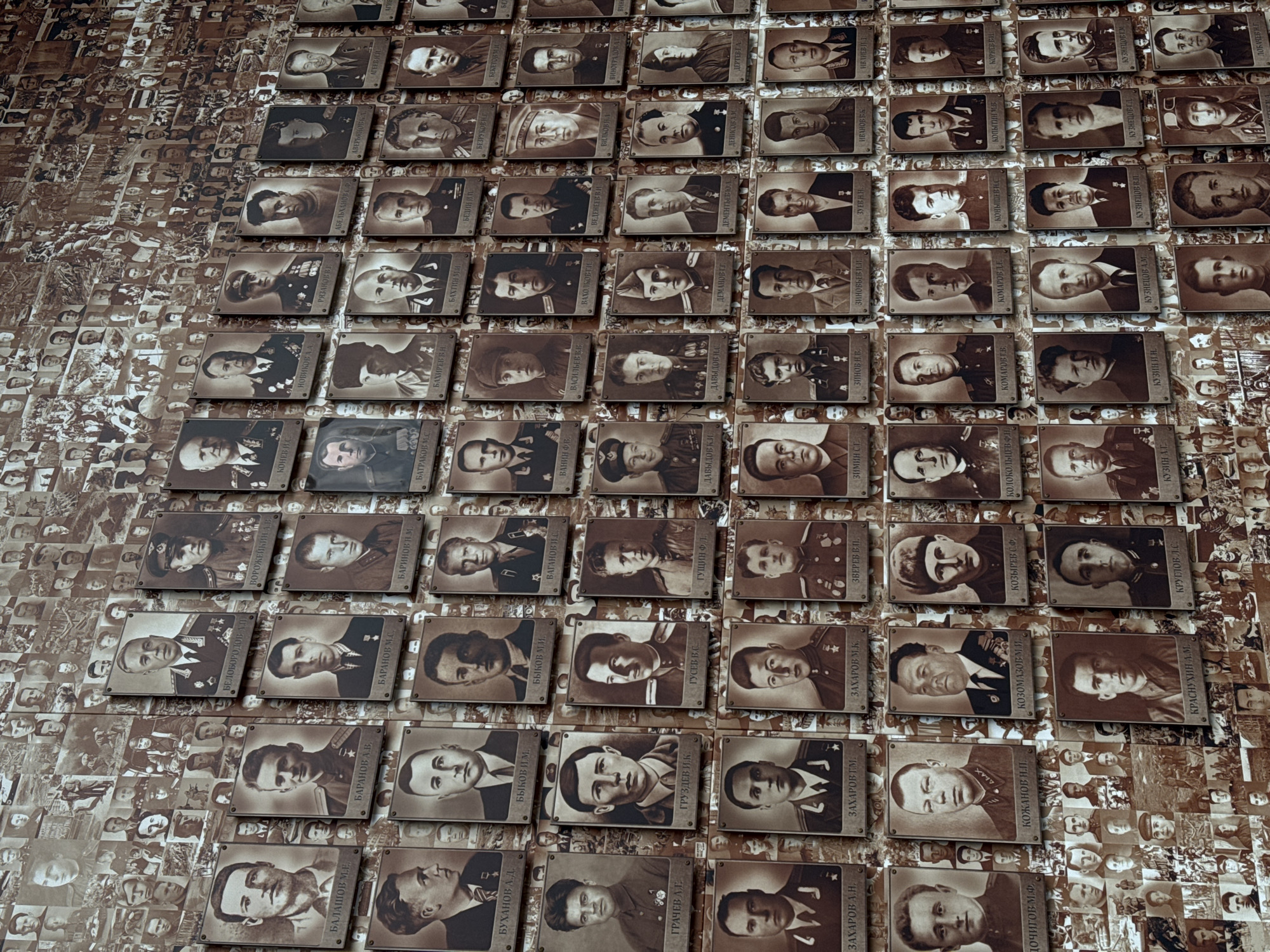 The image shows a wall covered with numerous small, sepia-toned portrait plaques. Each plaque displays a photograph of a person, presumably a soldier or historical figure, along with a name written in Cyrillic script. The plaques are arranged in a grid-like pattern, but the arrangement appears slightly irregular. The background behind the plaques is filled with smaller, faded images of people and events, creating a dense and layered visual effect. The overall impression is that of a memorial or tribute to individuals associated with military service or historical significance. The location is presumably in Nizhny Novgorod, Russia, but this cannot be definitively confirmed from the image alone.