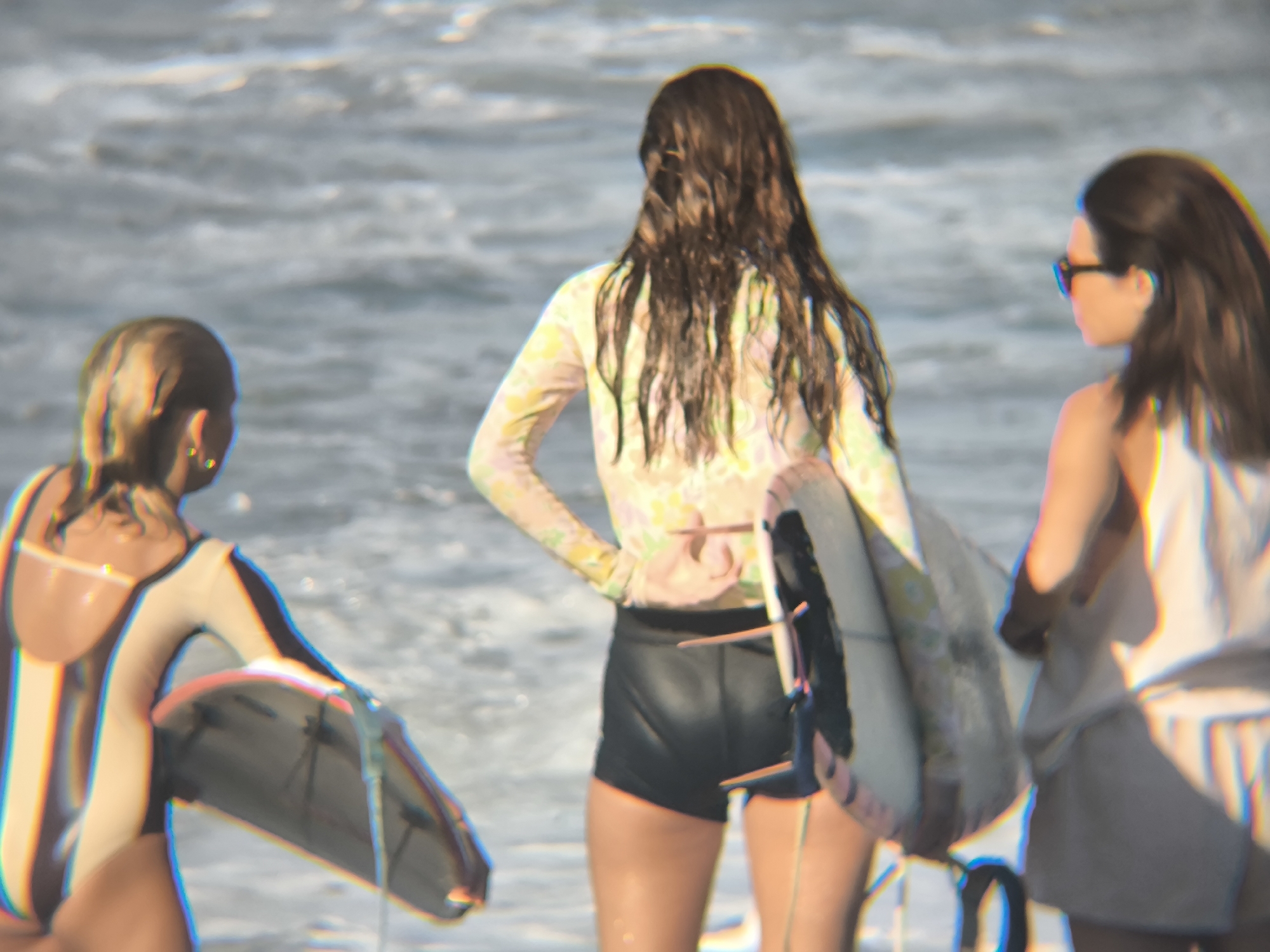 The image shows three women standing on a beach, presumably about to surf. The woman in the center has her back to the viewer and is wearing a long-sleeved rashguard with a floral pattern and black shorts. She is holding a surfboard. The woman on the left is wearing a one-piece swimsuit with dark vertical stripes and carrying a surfboard. The woman on the right is wearing sunglasses, a white tank top, and a grey skirt or shorts. She is also holding a surfboard. They are standing in shallow water near the shoreline, with the ocean waves visible in the background. While the image does not explicitly state the location, the prompt provides it as North Kuta, Indonesia.